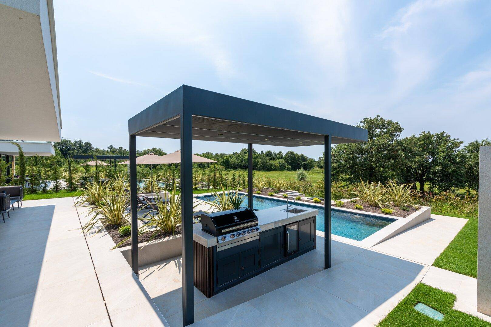 Moderne Außenküche mit Grill und Spüle neben dem Poolbereich unter einem schattenspendenden Vordach.