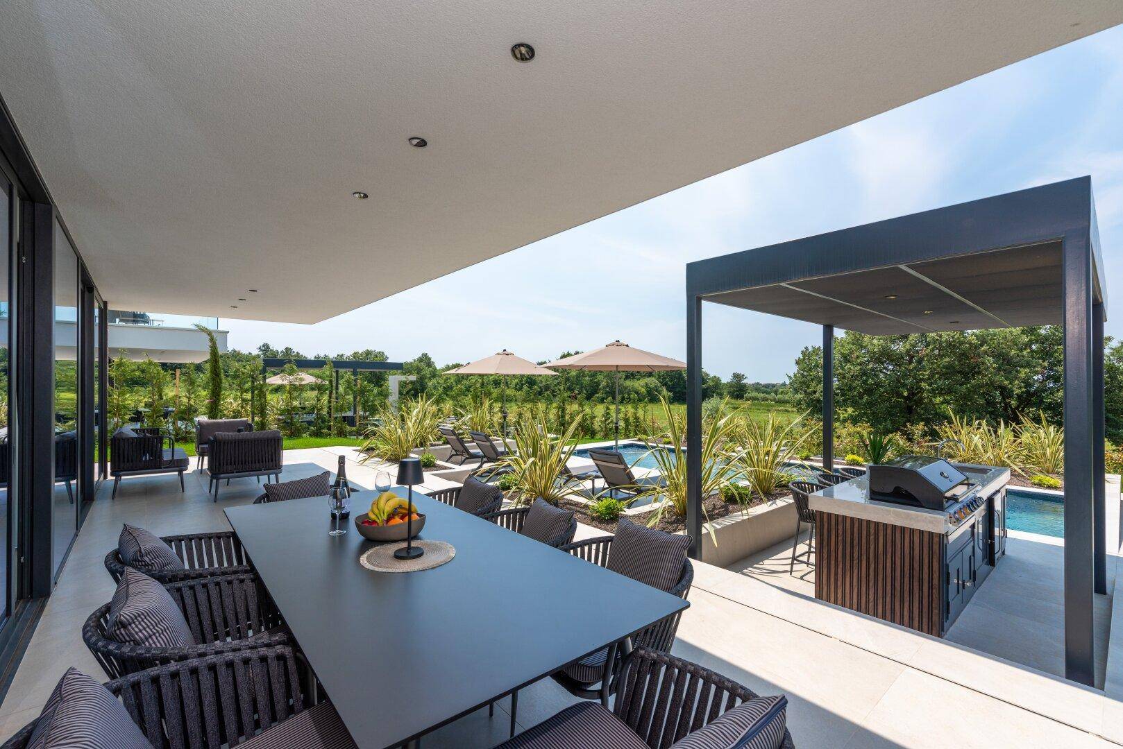 Elegante überdachte Terrasse mit großem Esstisch und Blick auf den Pool und die Außenküche.