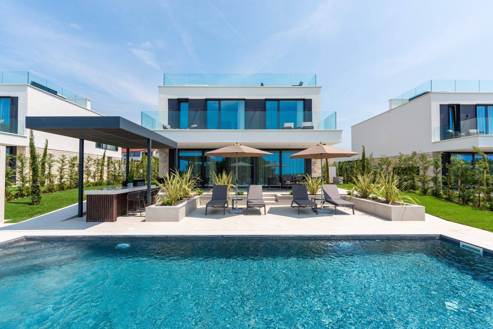 Moderne Villa mit großem Pool, Liegebereich und Glasbalkonen unter strahlend blauem Himmel.