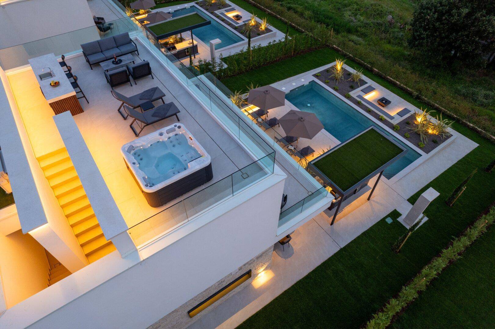 Beleuchtete Dachterrasse mit Whirlpool und Außenküche, sowie Poolbereich im Garten bei Dämmerung.