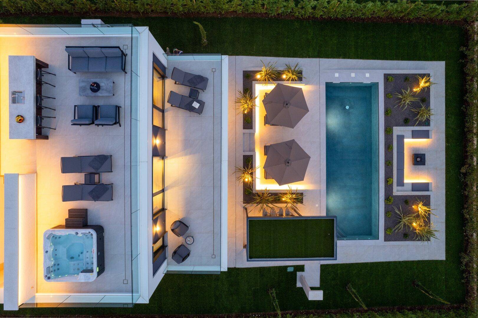 Vogelperspektive auf eine moderne Villa mit beleuchtetem Pool, Dachterrasse und Whirlpool bei Nacht.