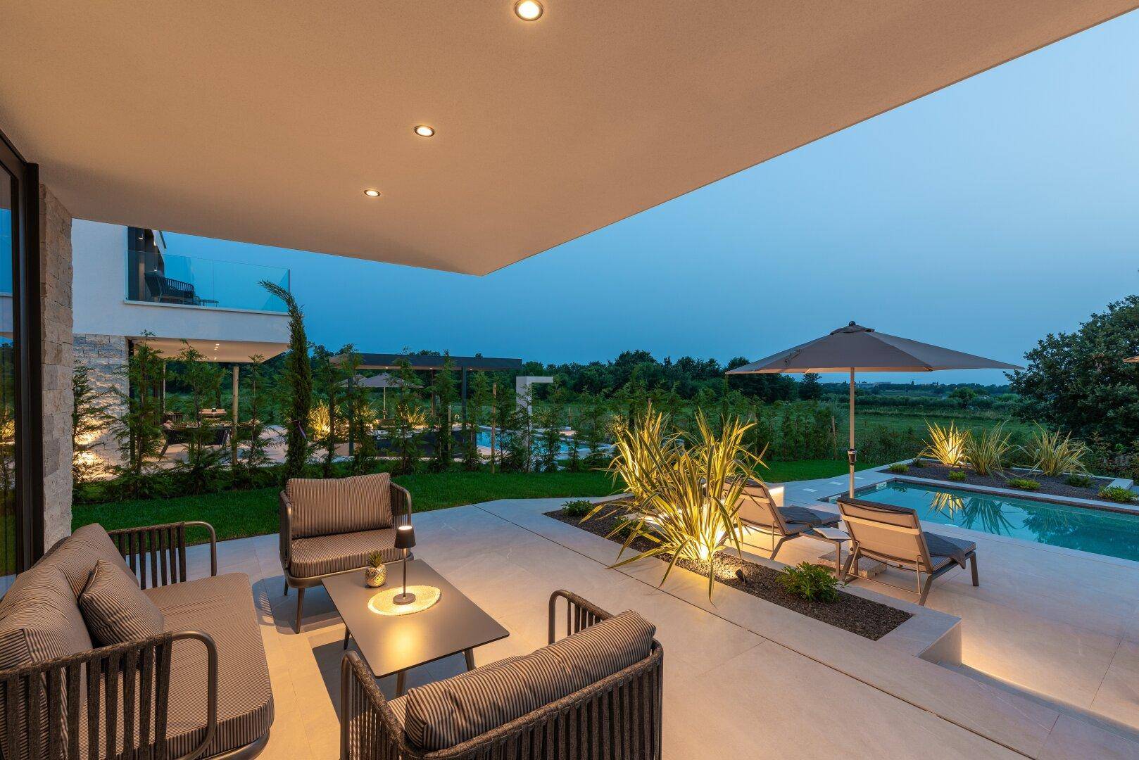 Gemütliche überdachte Terrasse mit Lounge-Möbeln und stimmungsvoller Beleuchtung bei Dämmerung.