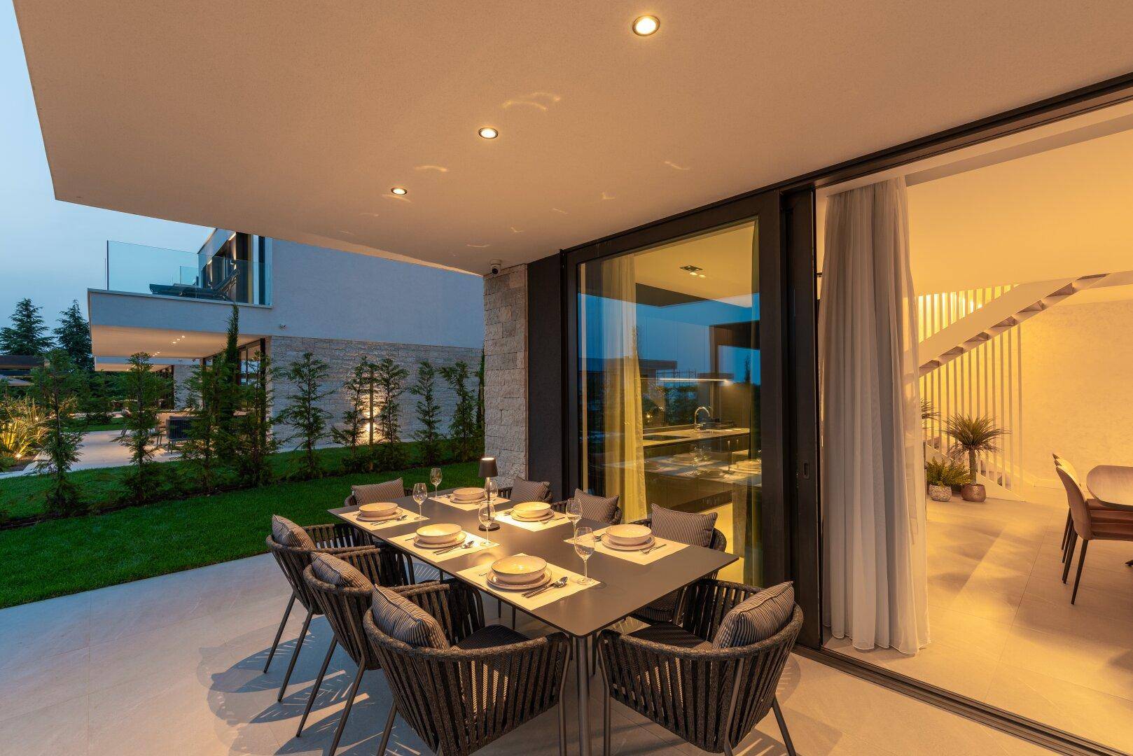 Elegante überdachte Terrasse mit Esstisch und Blick auf den beleuchteten Garten bei Dämmerung.