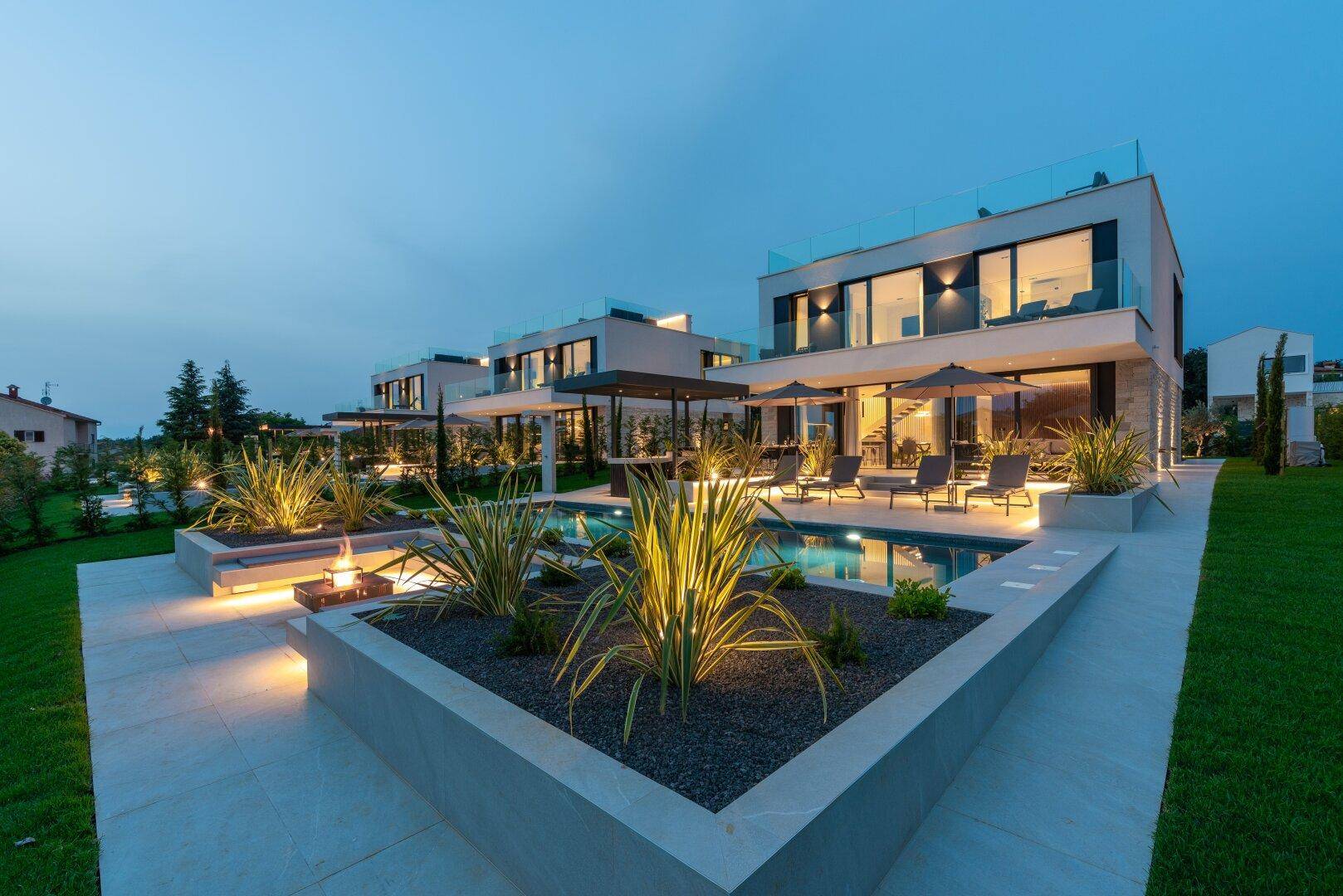 Moderne Villa mit beleuchtetem Pool, Feuerstelle und stilvoller Außenbeleuchtung bei Dämmerung.