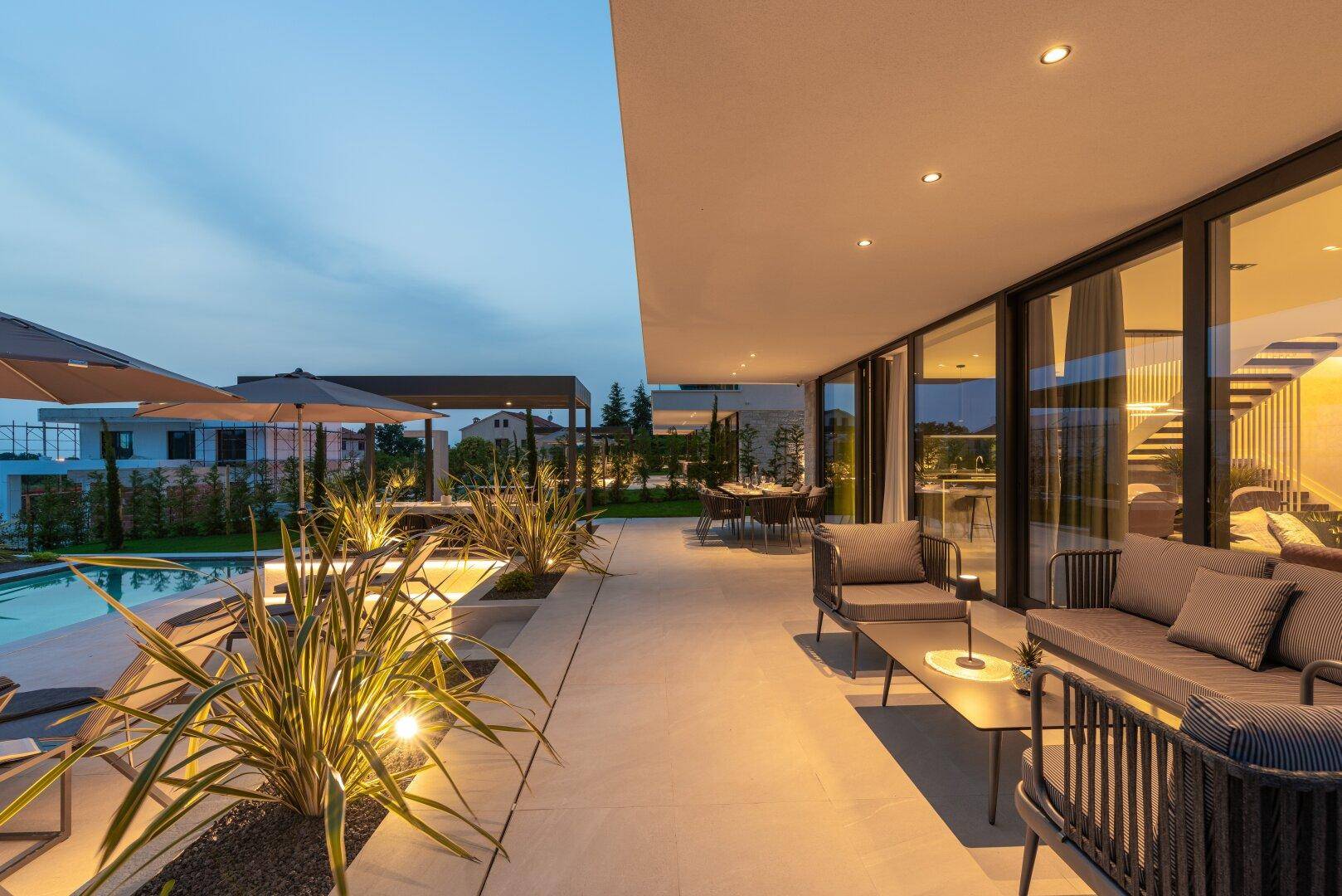 Stilvolle überdachte Terrasse mit Lounge-Möbeln und Blick auf den beleuchteten Poolbereich bei Dämmerung.