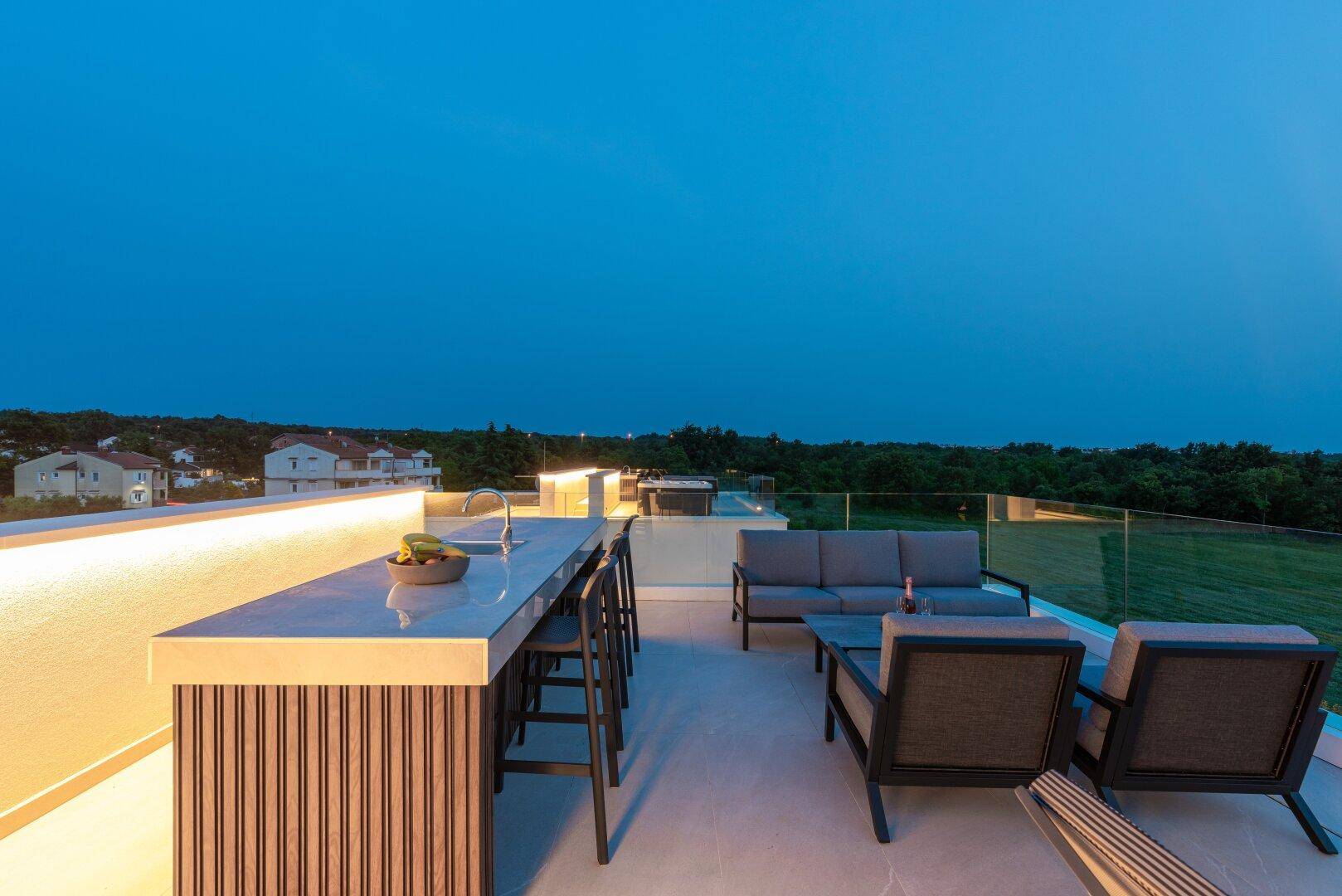 Moderne Dachterrasse mit beleuchteter Außenküche, Barhockern und Lounge-Bereich bei Dämmerung.