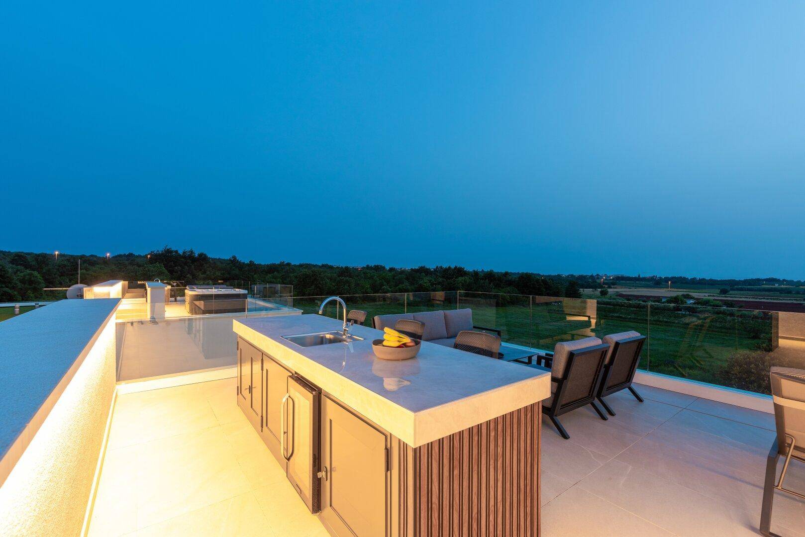 Stilvolle Dachterrasse mit beleuchteter Außenküche und weitem Blick über die Umgebung bei Dämmerung.