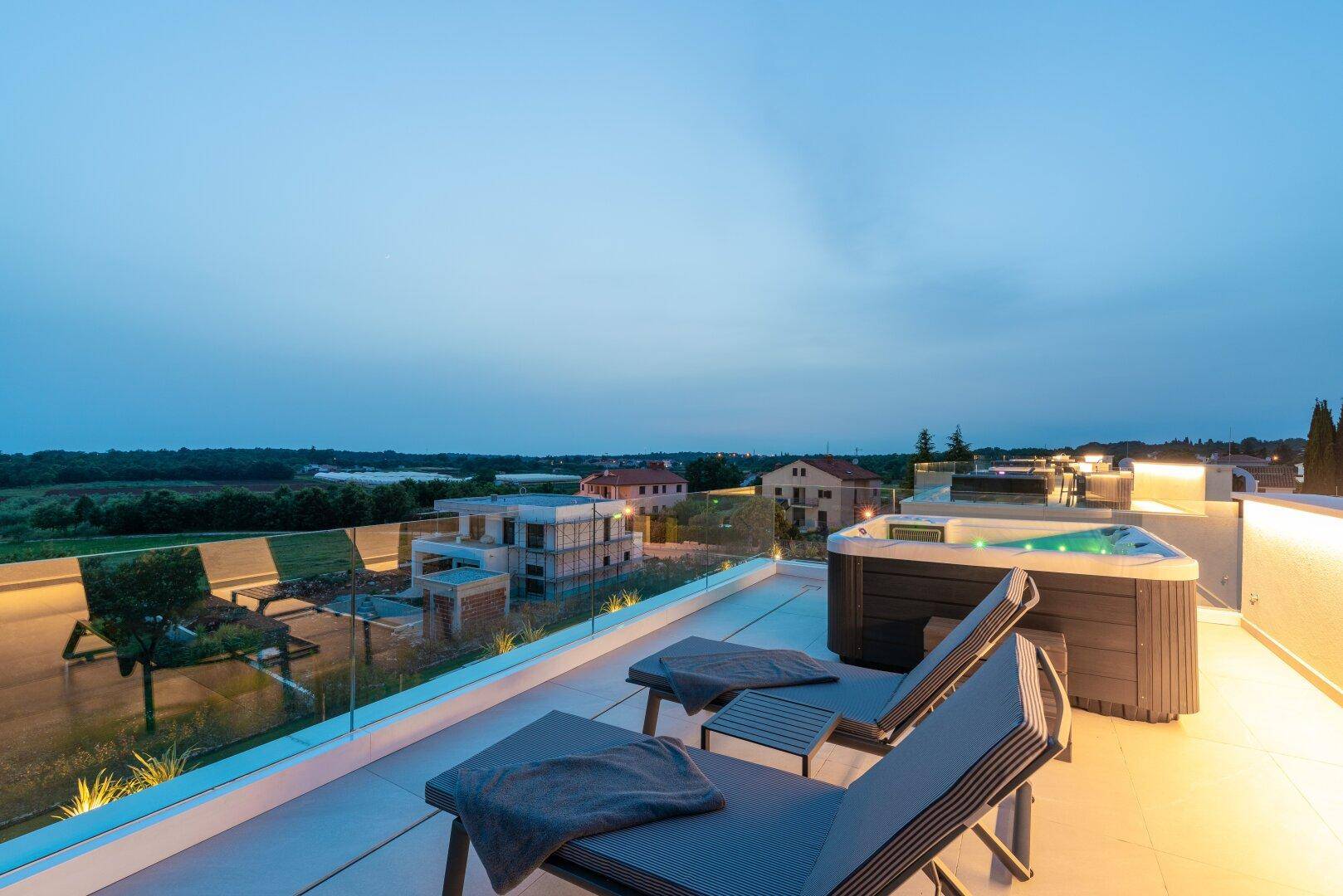 Entspannende Dachterrasse mit Sonnenliegen und Whirlpool, umgeben von einer Glasbrüstung bei Dämmerung.