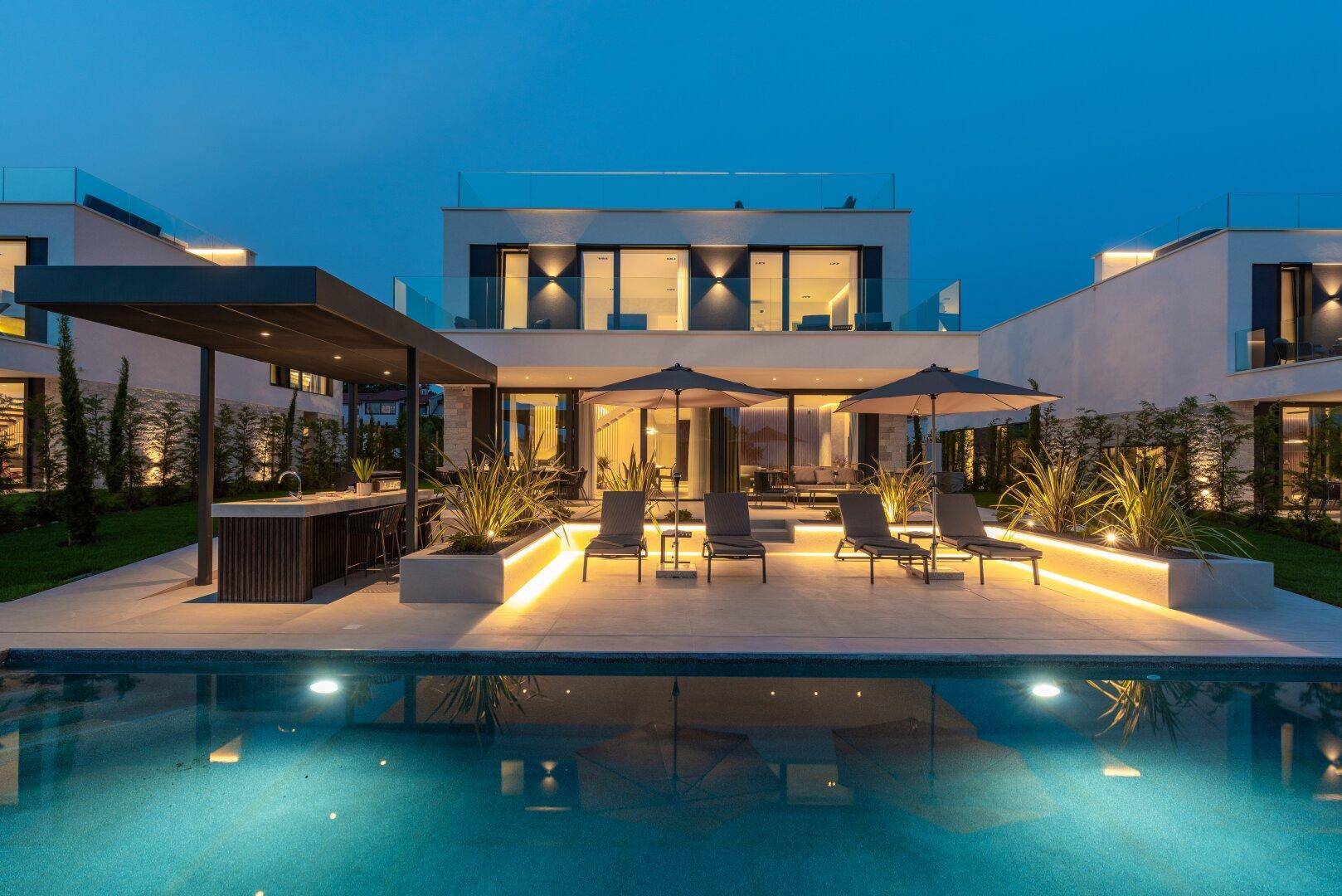 Elegante Villa mit beleuchtetem Pool und Liegebereich, umgeben von stilvoller Außenbeleuchtung bei Dämmerung.