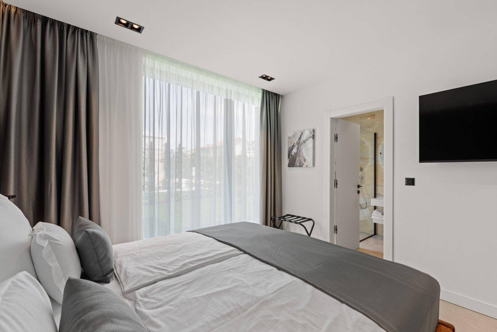 Helles Schlafzimmer mit Doppelbett, Fernseher und Zugang zu einem modernen Badezimmer.