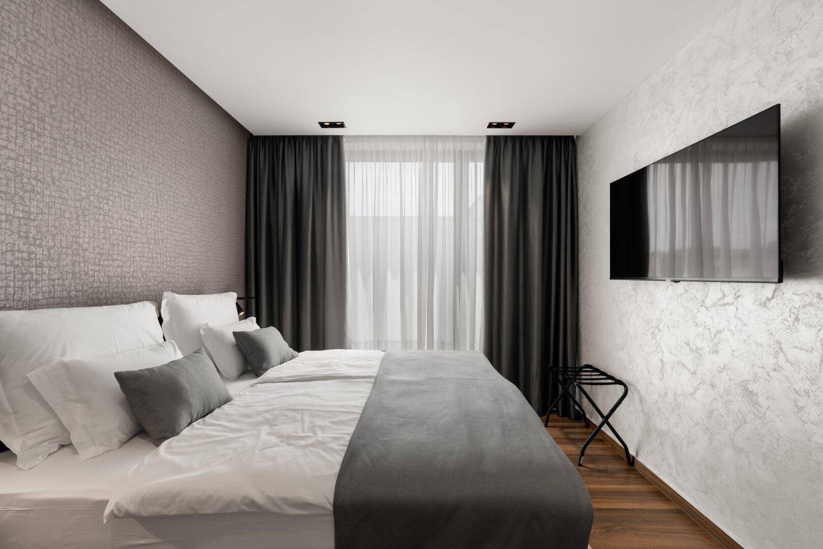 Helles Schlafzimmer mit Doppelbett, Fernseher und großen Fenstern mit Blick nach draußen.