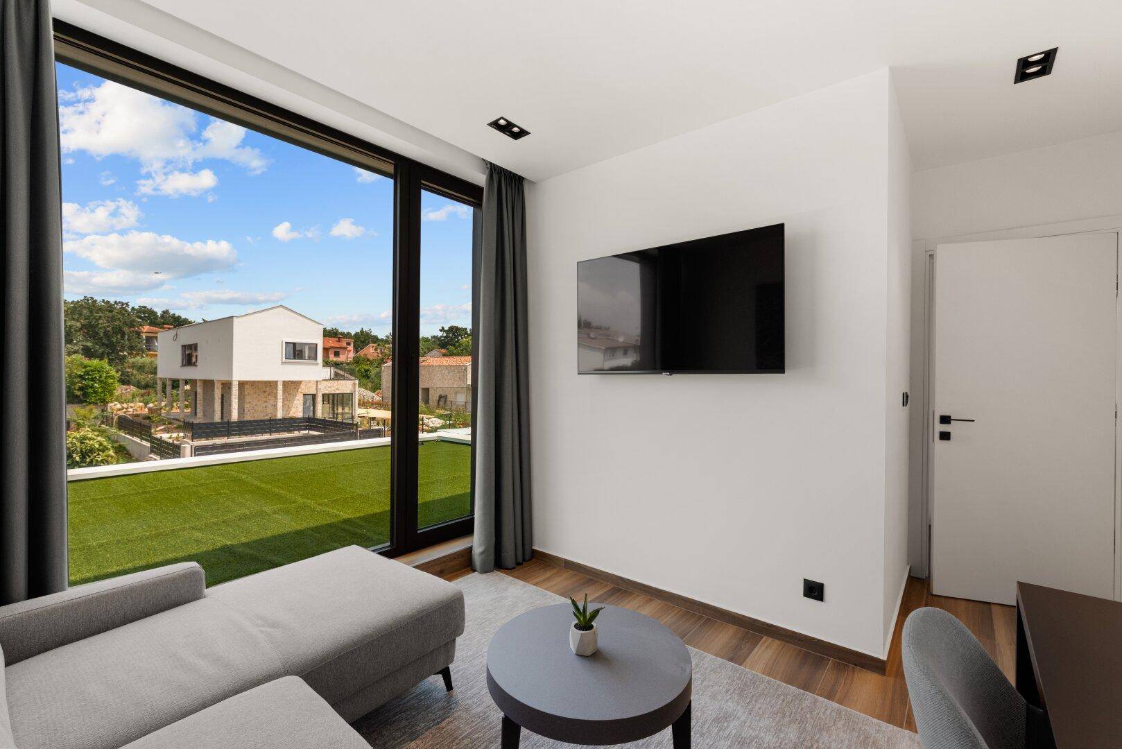 Modernes Wohnzimmer mit Ecksofa, Fernseher und großen Fenstern mit Blick auf die Umgebung.