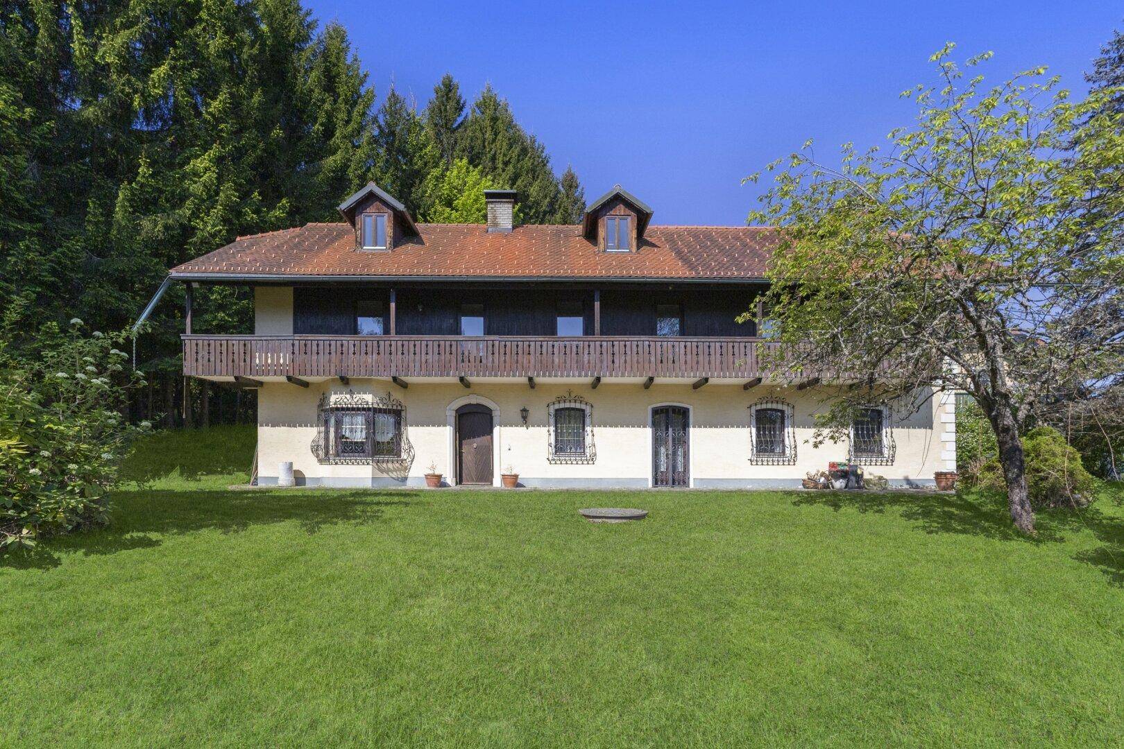 Frontansicht des Landhauses mit traditionellen Fenstern, Holzbalkon und weitläufiger Rasenfläche.