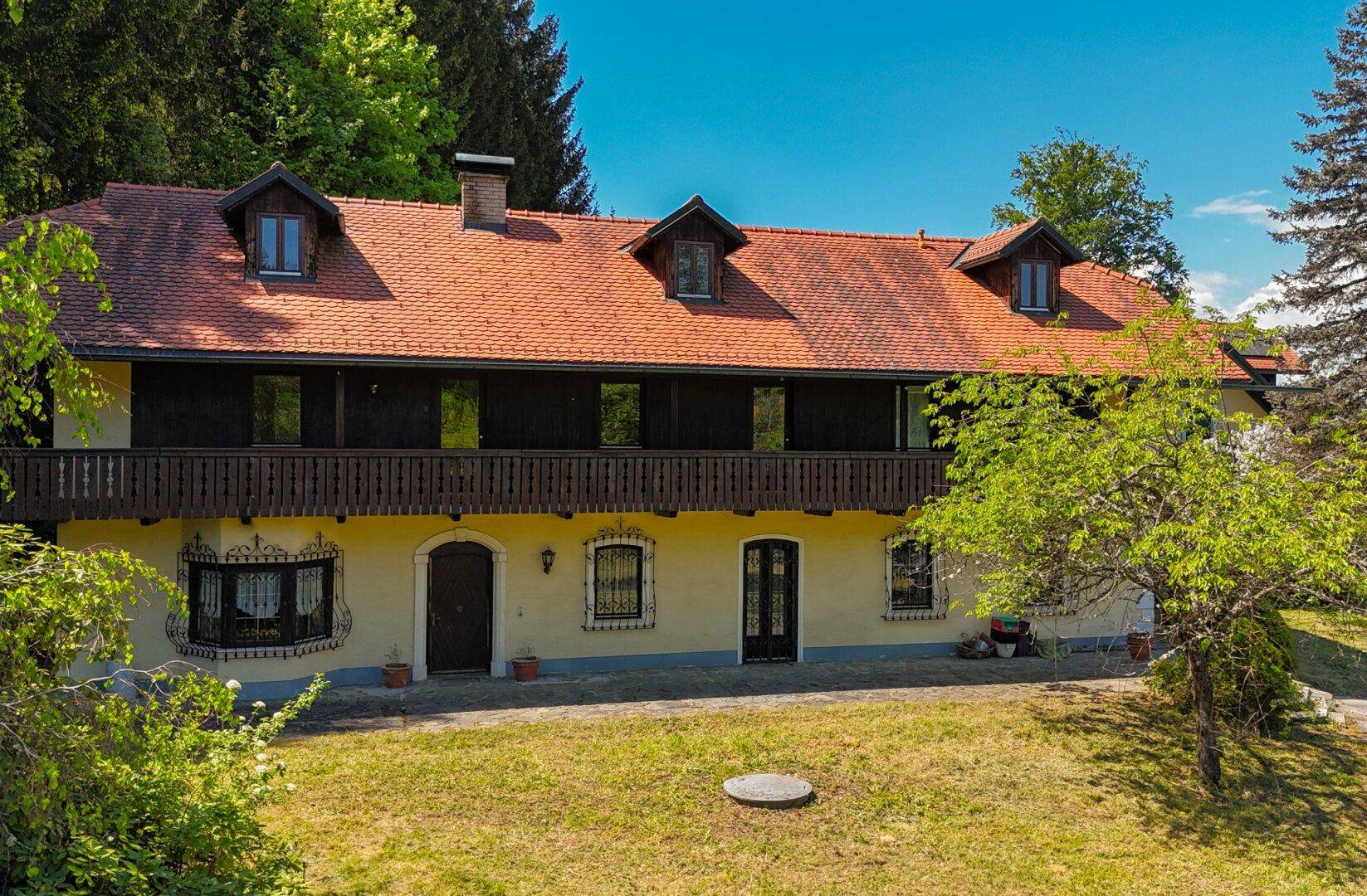 Traditionelles Landhaus mit rotem Ziegeldach, Holzbalkonen und großem Grundstück in grüner Umgebung.