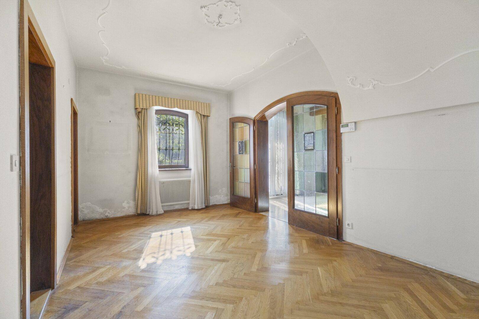 Geräumiges Wohnzimmer mit Fischgrätparkett, hohen Decken und eleganten Flügeltüren.