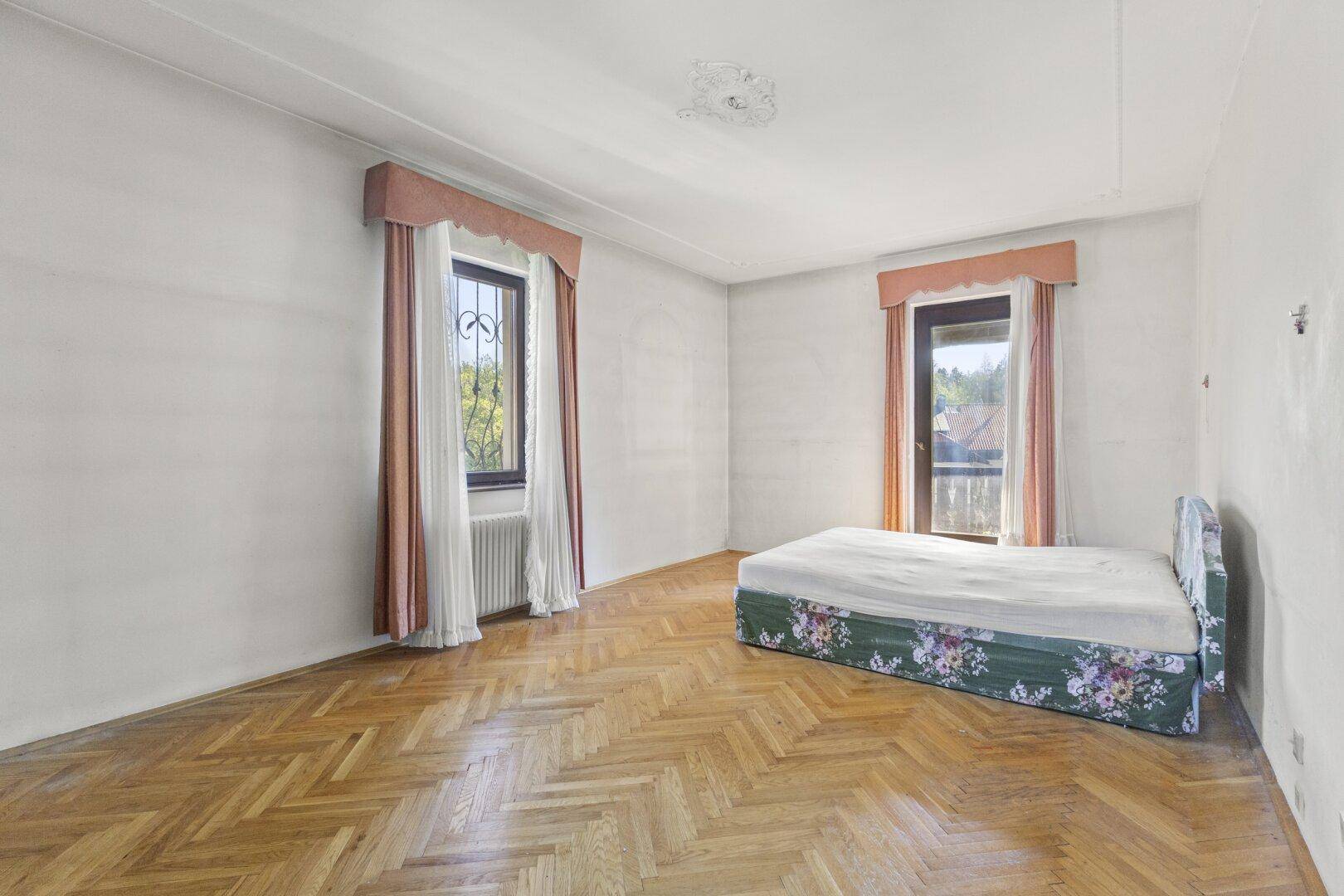 Geräumiges Schlafzimmer mit Fischgrätparkett, hohen Decken und zwei Fenstern.
