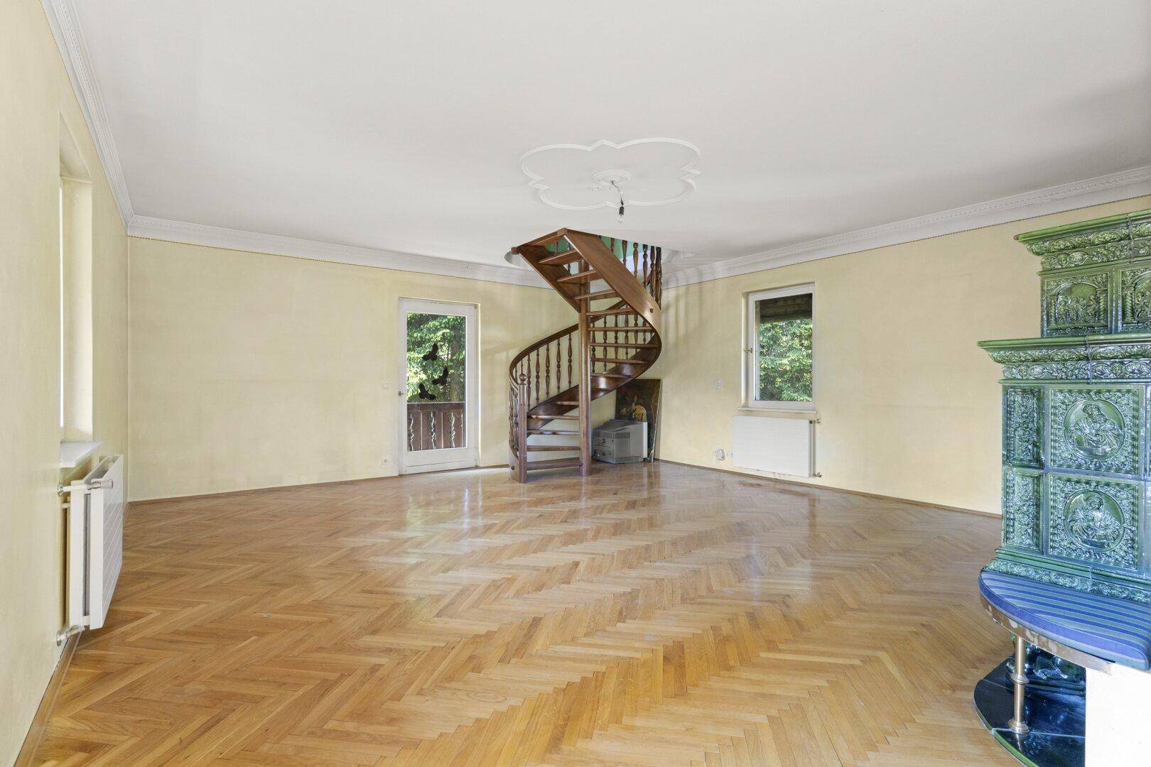 Helles Wohnzimmer mit Fischgrätparkett, grünem Kachelofen und Wendeltreppe.