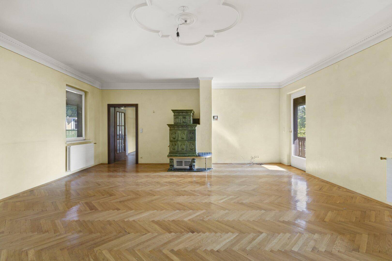 Geräumiges Wohnzimmer mit Fischgrätparkett, grünem Kachelofen und Zugang zum Balkon.