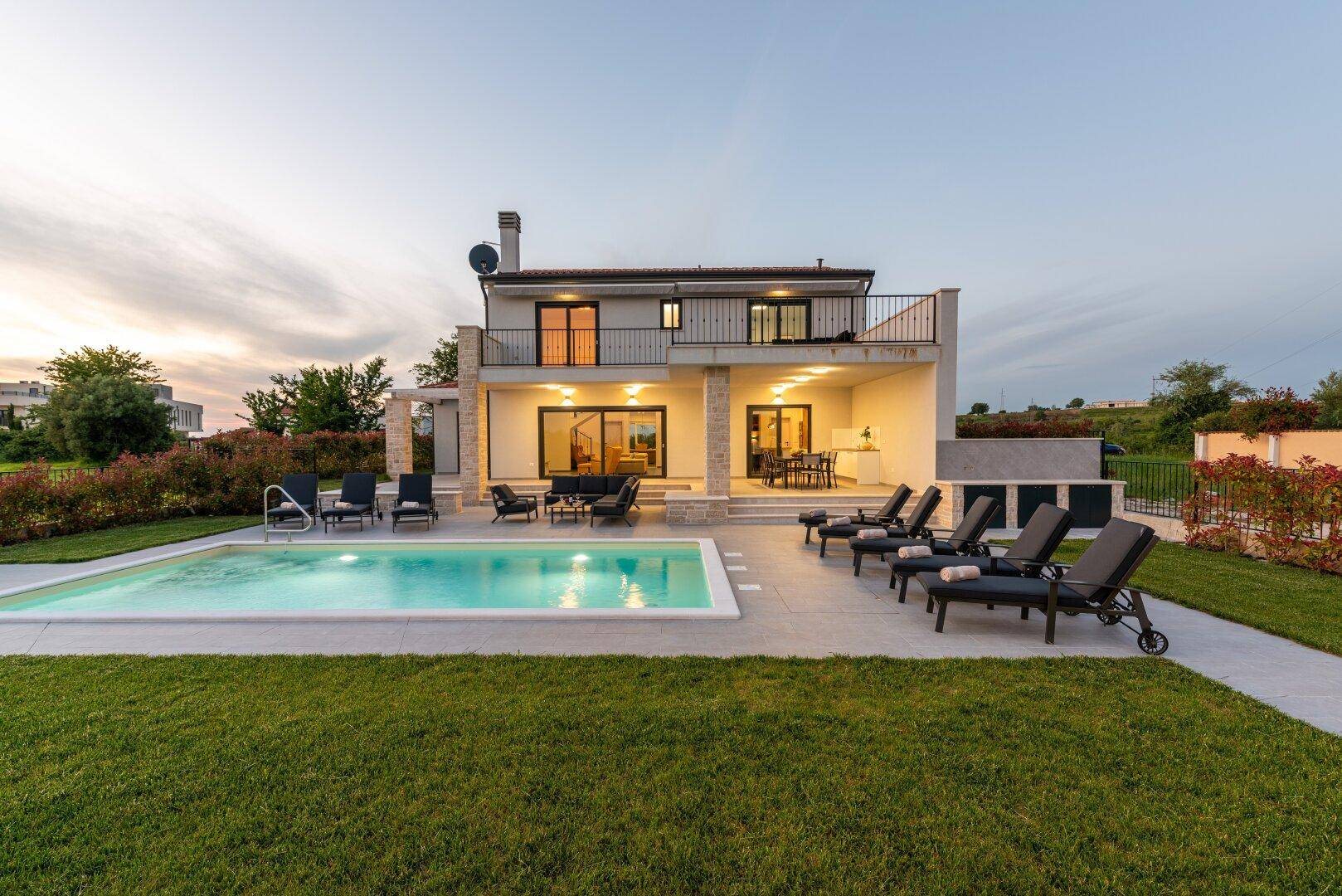 Abendliche Ansicht einer modernen Villa mit beleuchtetem Pool und großzügiger Terrasse mit Sitzgelegenheiten.