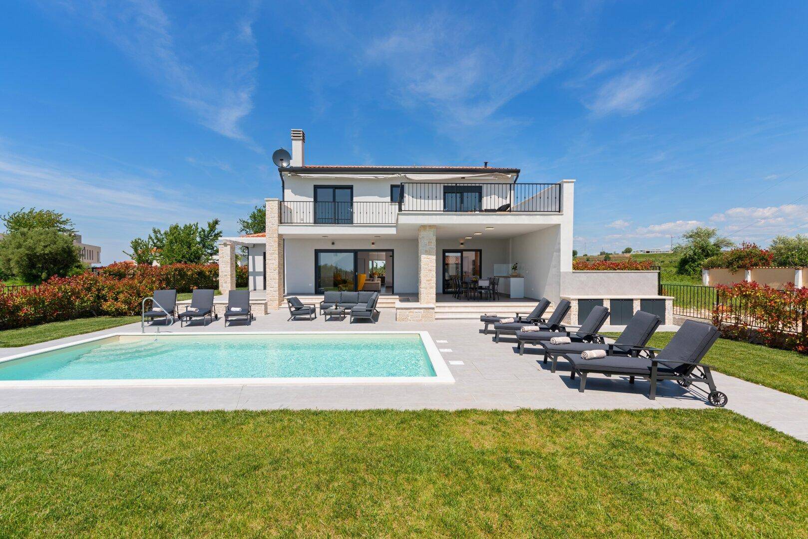 Sonnige Ansicht einer modernen Villa mit großem Pool und weitläufiger Terrasse mit Liegestühlen.