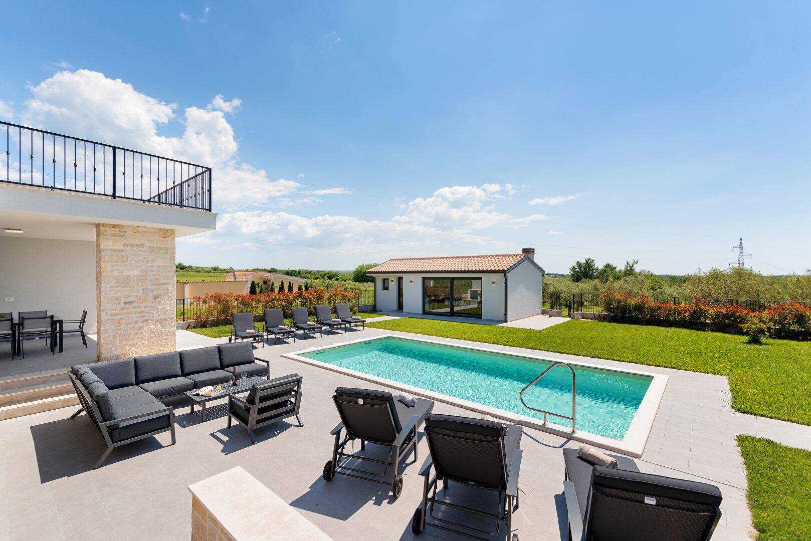 Panoramablick auf den Poolbereich mit Lounge-Möbeln, Liegestühlen und dem Nebengebäude im Grünen.