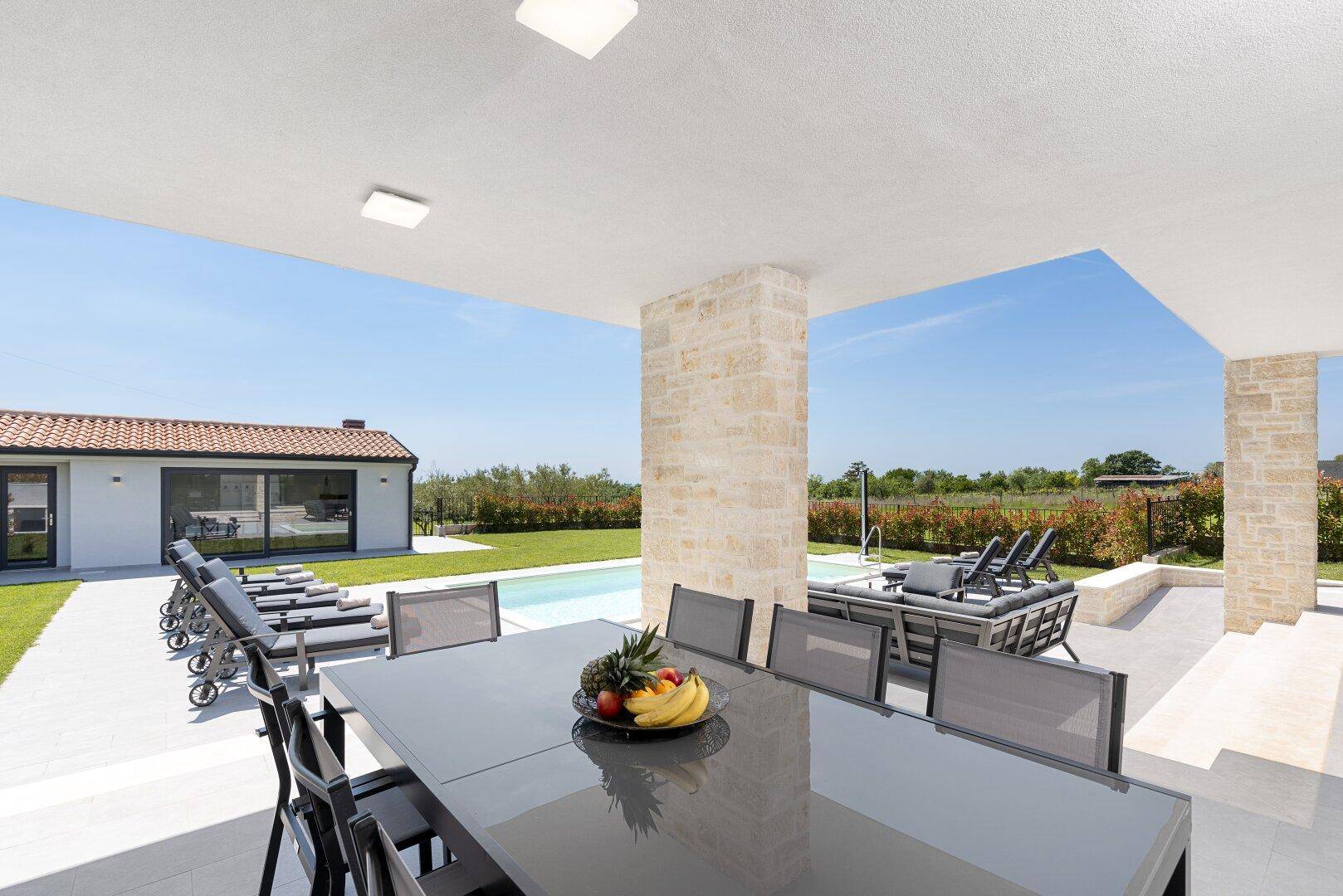 Überdachter Essbereich im Freien mit großem Tisch und Blick auf den Pool und die Landschaft.