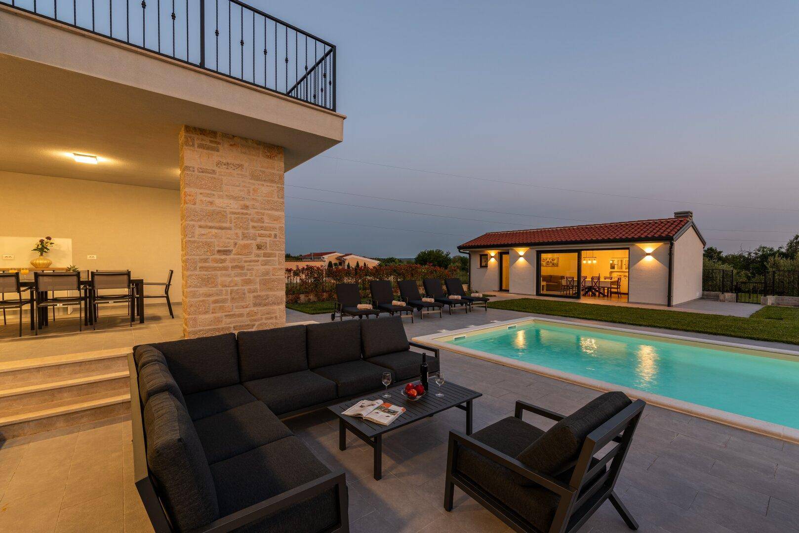 Gemütlicher Lounge-Bereich im Freien mit Blick auf den beleuchteten Pool und das Nebengebäude bei Dämmerung.
