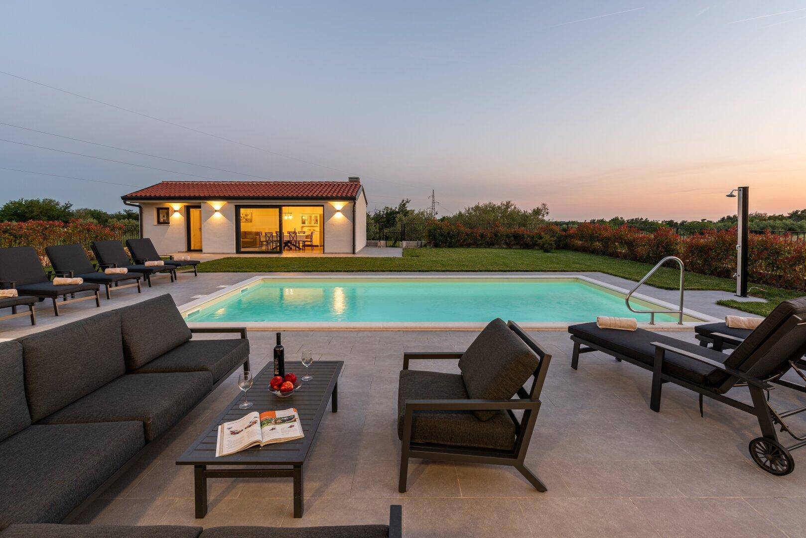 Entspannter Lounge-Bereich im Freien mit Blick auf den beleuchteten Pool und das Nebengebäude bei Dämmerung.