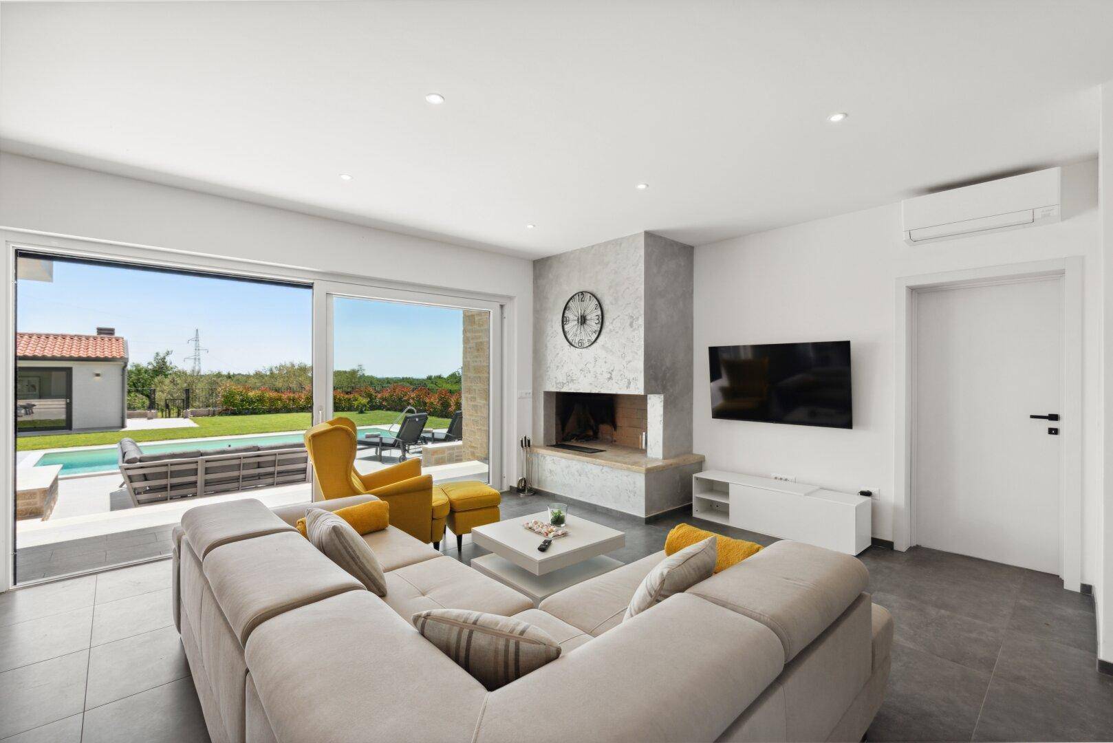Helles Wohnzimmer mit großem Sofa, modernem Kamin und Zugang zur Terrasse mit Poolblick.
