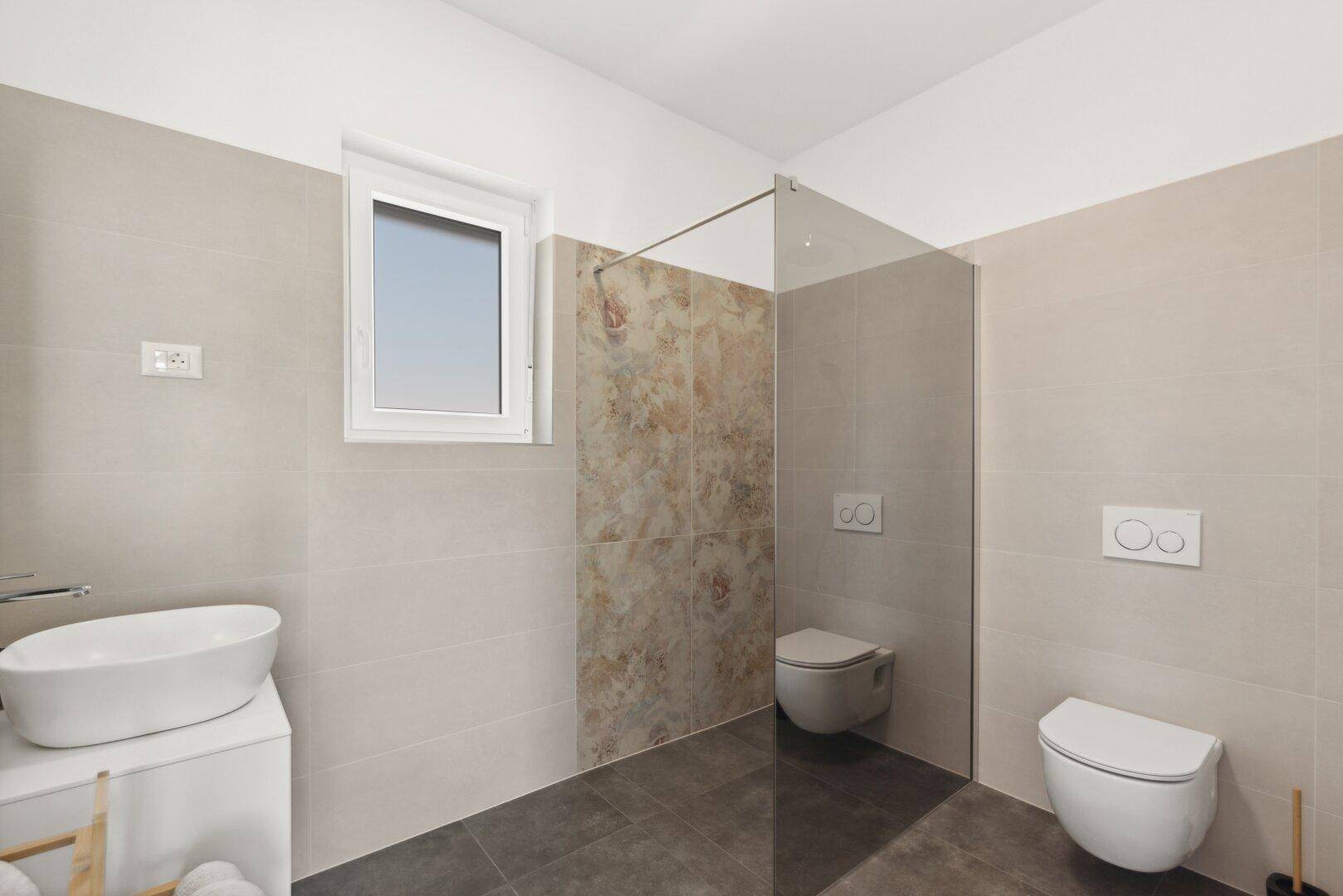 Helles Badezimmer mit Fenster, moderner Toilette und floral gemusterten Akzentfliesen an der Wand.