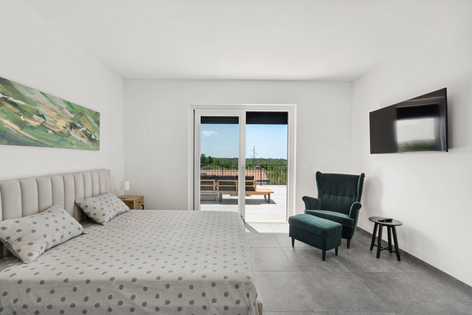 Stilvolles Schlafzimmer mit bequemen Sessel, Wandfernseher und Zugang zur sonnigen Terrasse.