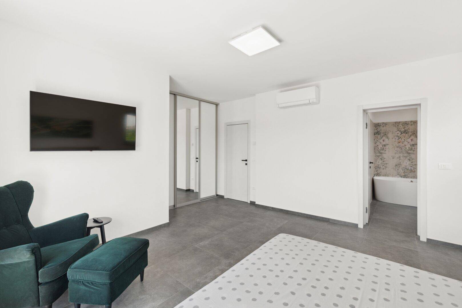 Helles Schlafzimmer mit Fliesenboden, Fernseher und Zugang zu einem modernen Badezimmer.