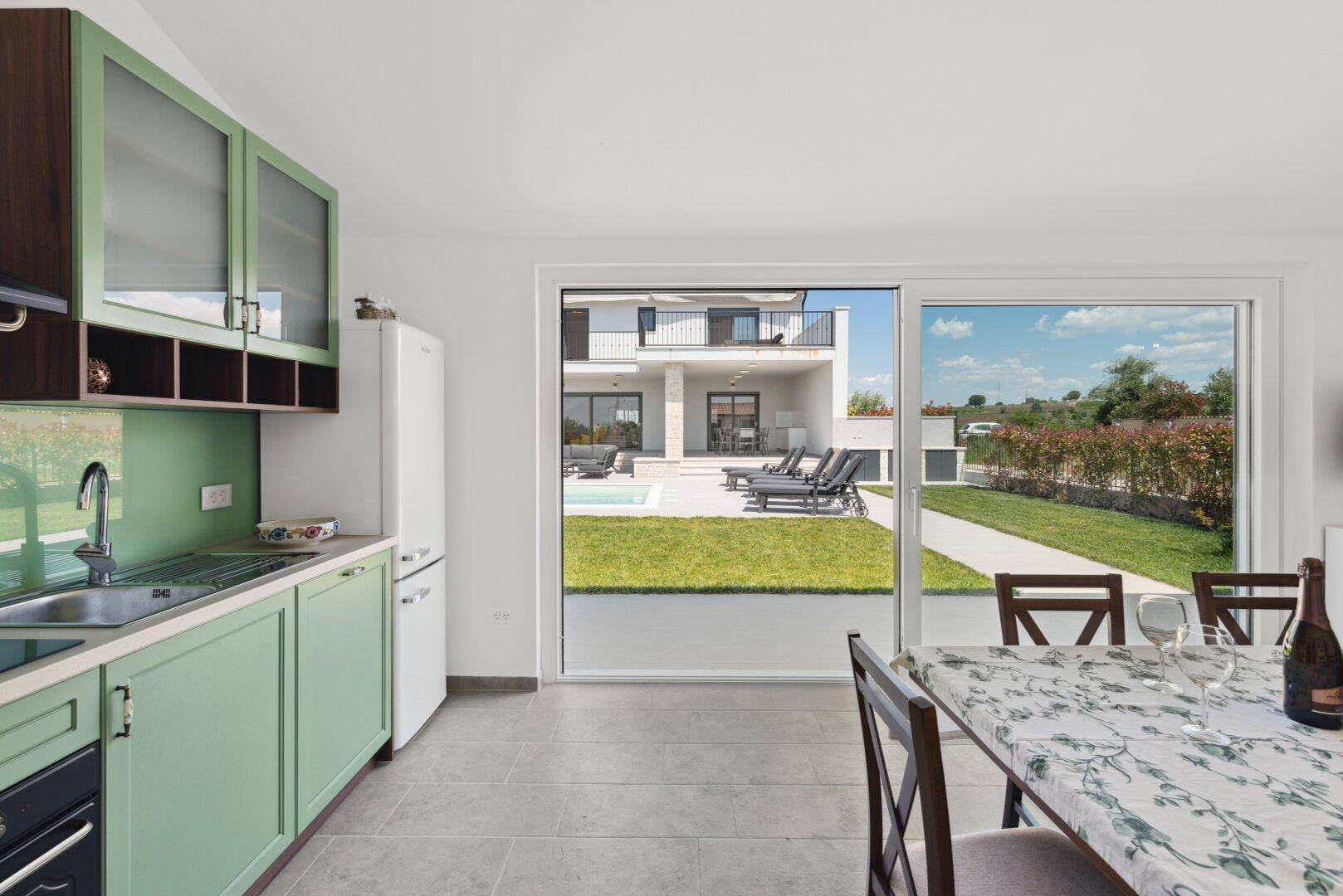 Moderne Küche mit grünen Schränken, Kühlschrank und großem Fenster zum Garten und Pool.