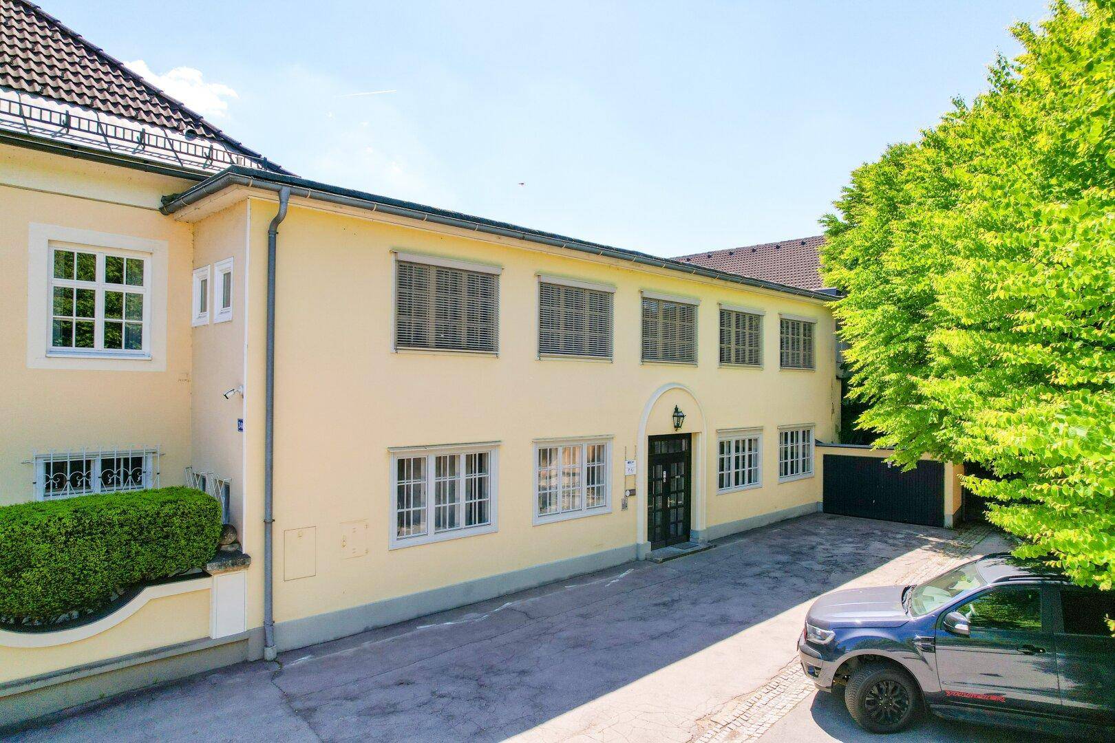 Ansicht der Gebäudefassade mit vielen Fenstern, Jalousien und einem gepflasterten Vorplatz.
