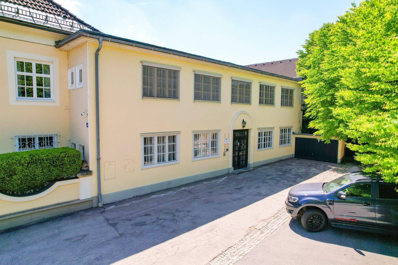 Gebäudefassade mit vielen Fenstern, Jalousien und einem gepflasterten Vorplatz mit parkendem Auto.