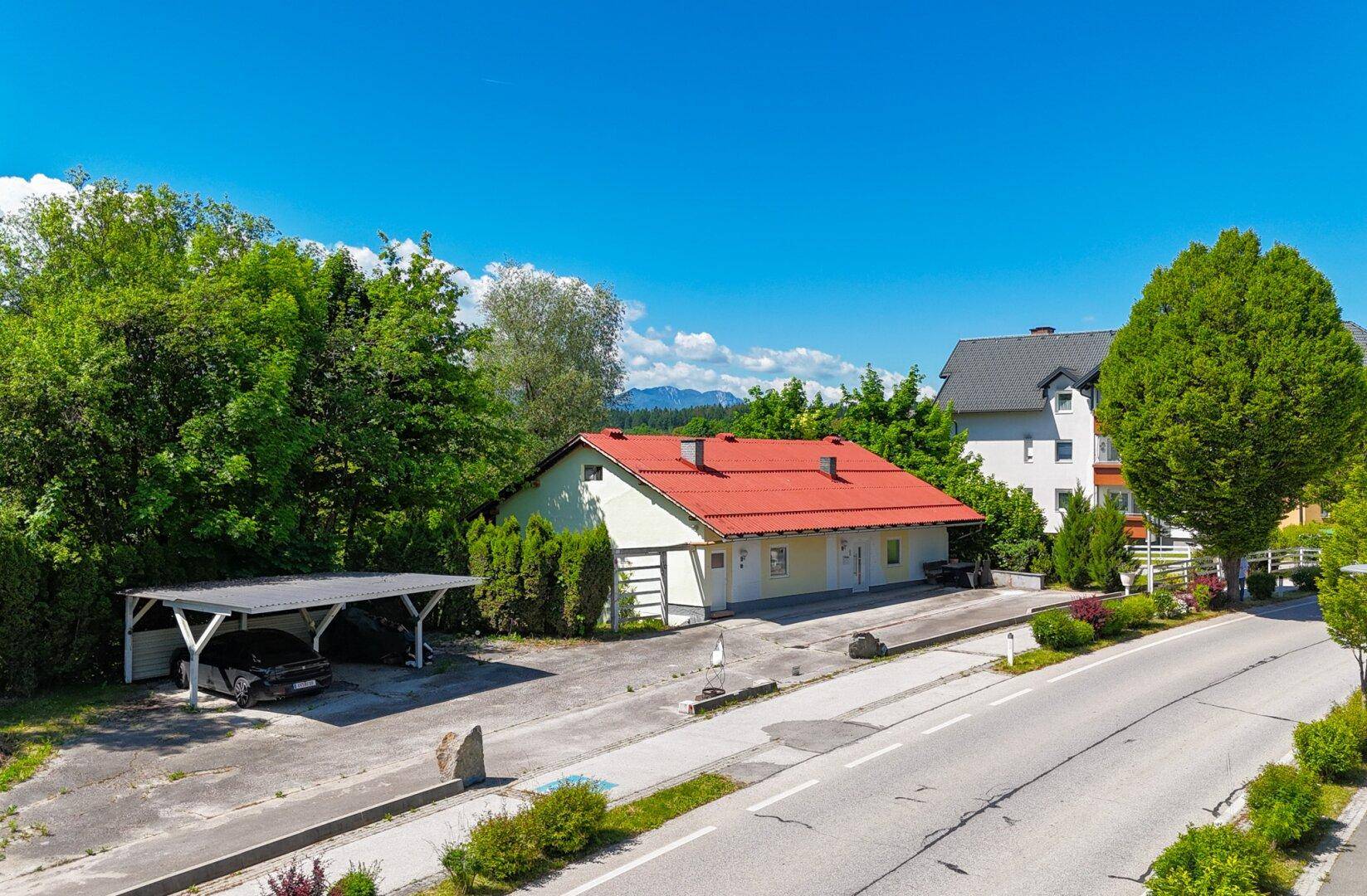 Wohnhaus mit Carport und Zufahrt, umgeben von Bäumen und mit Bergblick im Hintergrund.