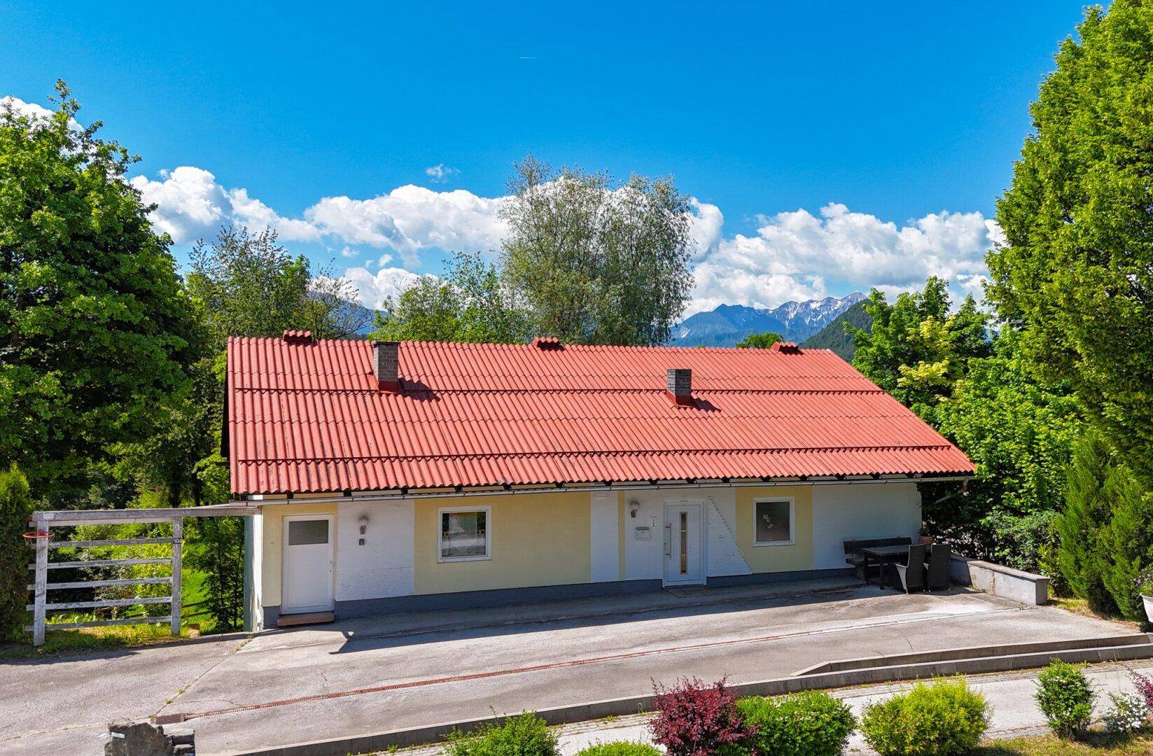 Vorderansicht des Hauses mit gelber Fassade, rotem Dach und Bergblick im Hintergrund.