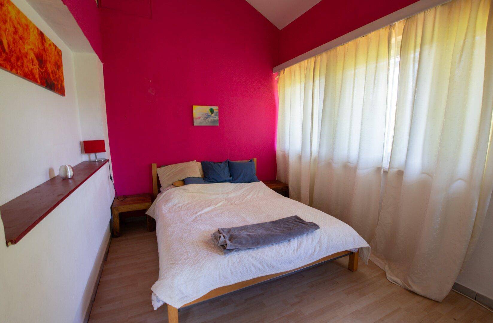 Schlafzimmer mit Doppelbett, Laminatboden und leuchtend pinker Akzentwand.