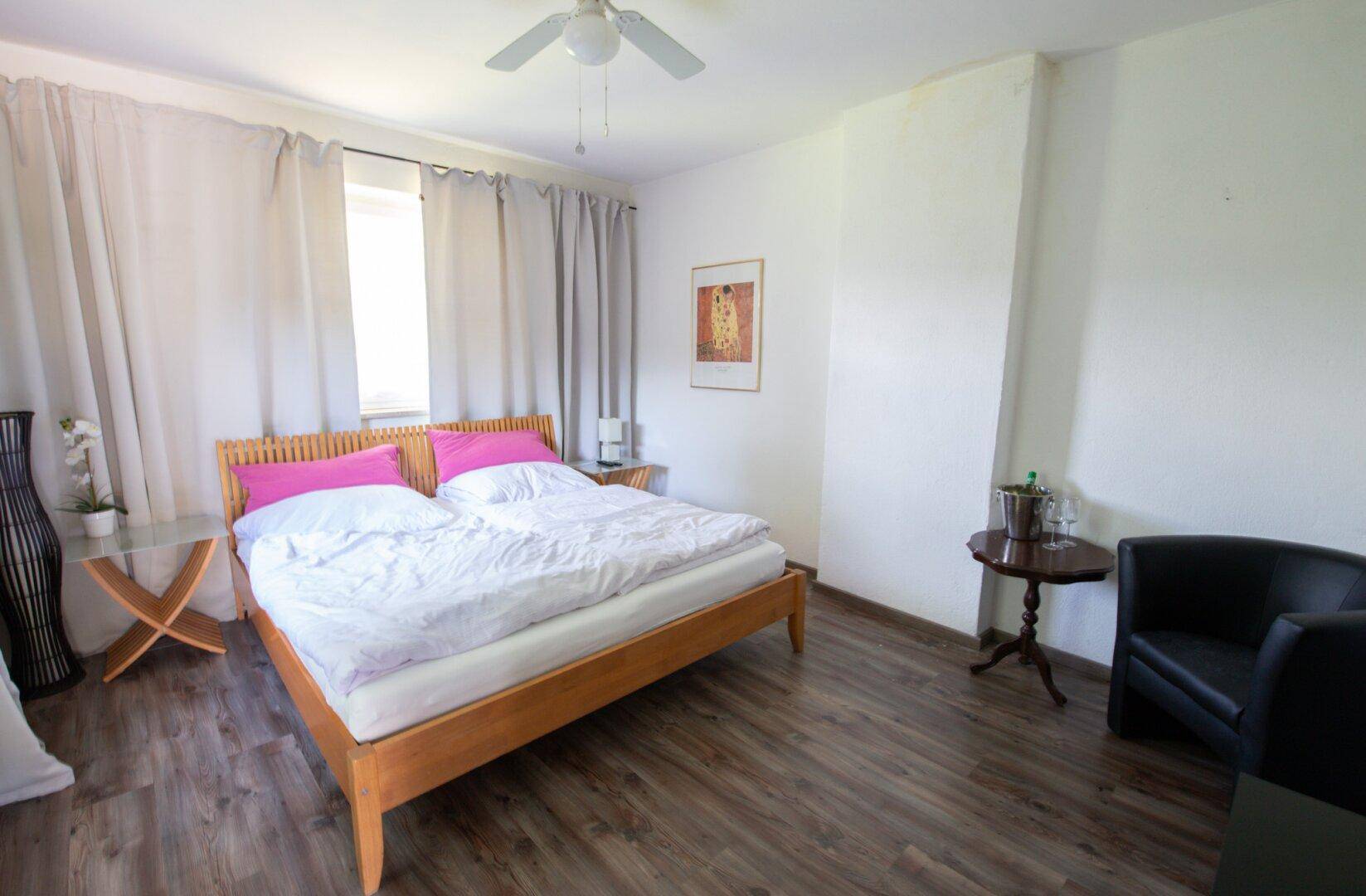 Schlafzimmer mit Doppelbett, Laminatboden, Deckenventilator und hellen Vorhängen.