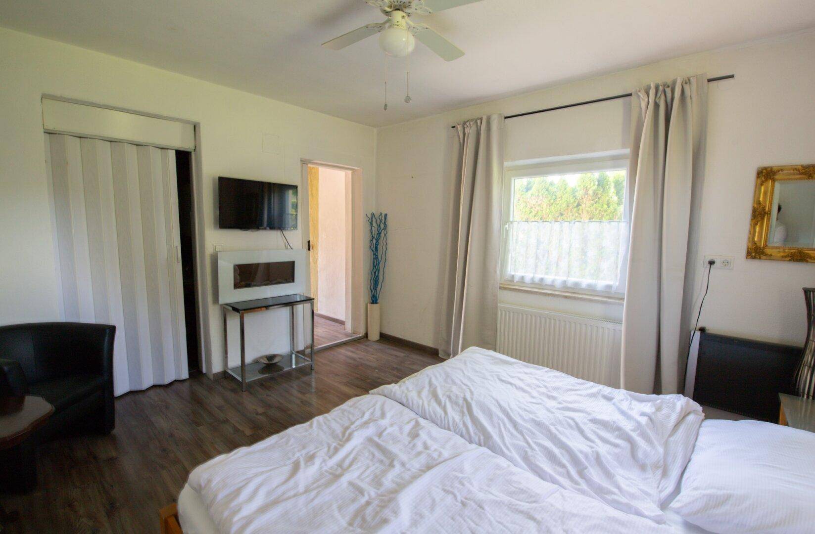Helles Schlafzimmer mit Doppelbett, Fenster und Fernseher an der Wand.