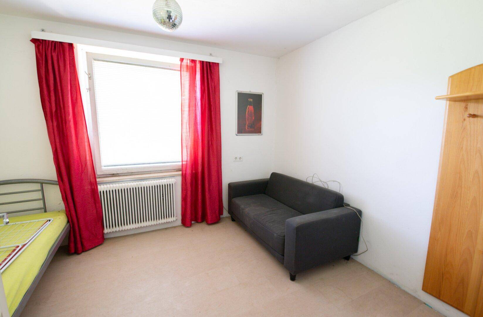 Helles Zimmer mit Einzelbett, Sofa und rotem Vorhang am Fenster.