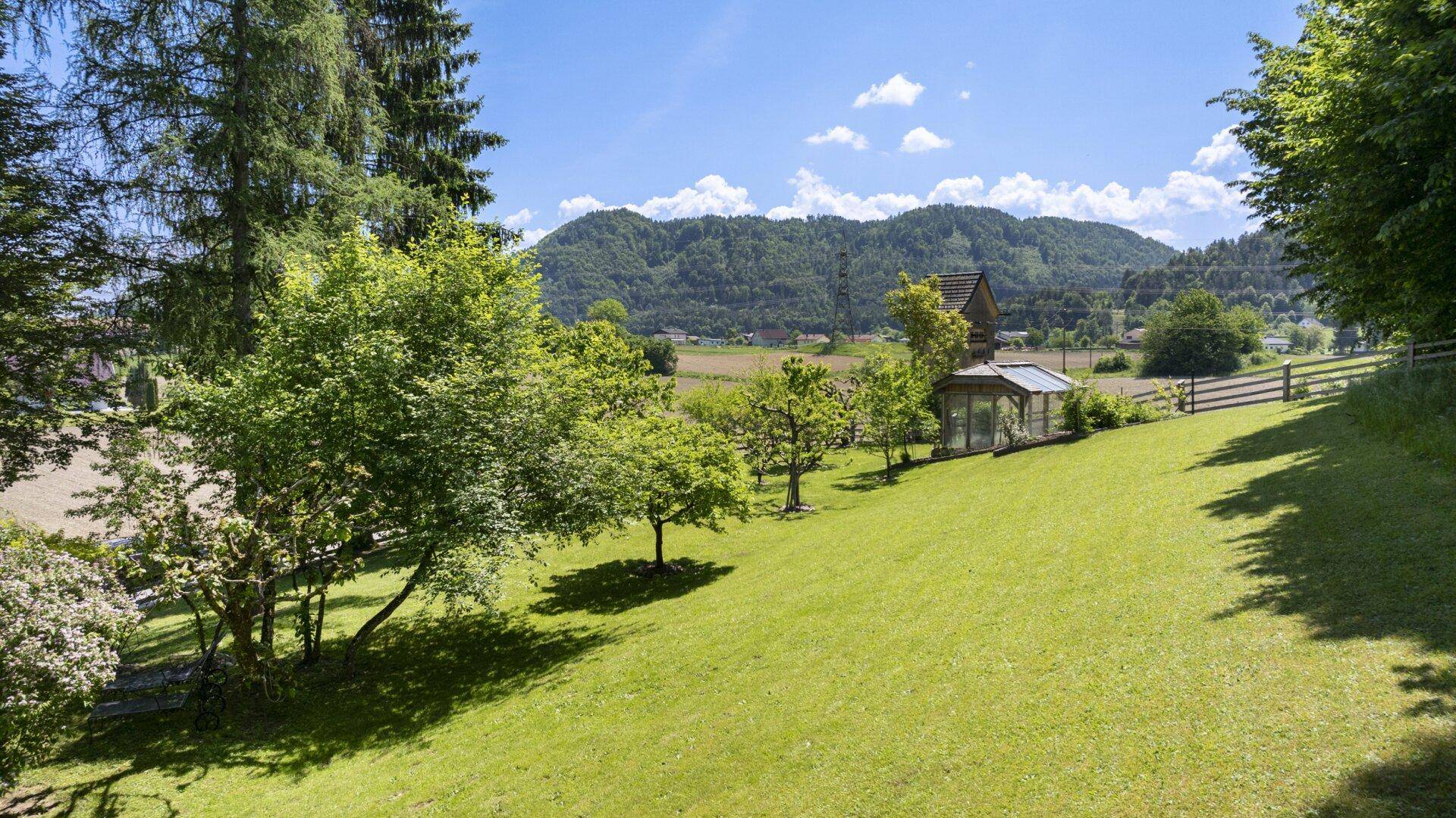 Weitläufiger grüner Garten mit sanften Hügeln und einem Gewächshaus, umgeben von Natur.