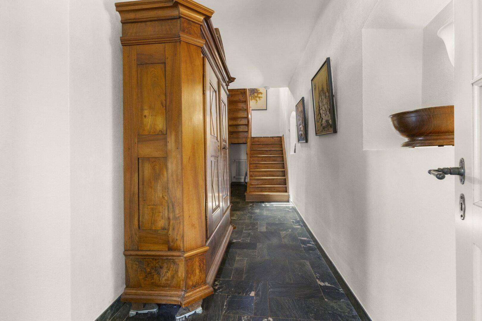 Langer Flur mit dunklem Steinboden, einem antiken Holzschrank und einer Holztreppe.