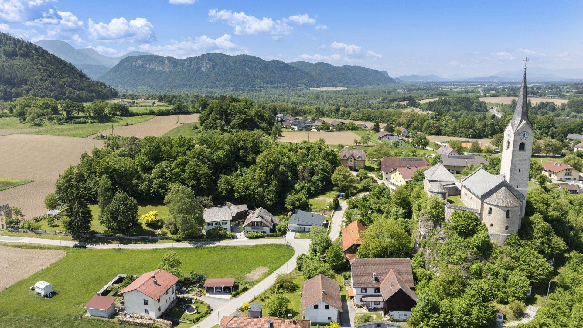 Luftaufnahme eines malerischen Dorfes mit Kirche, umgeben von grünen Feldern und Bergen unter blauem Himmel.