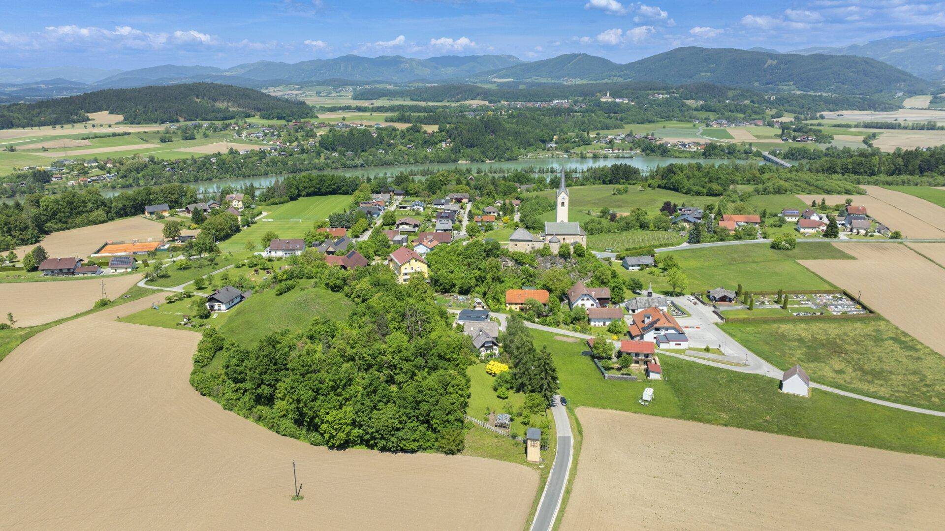 Weitläufige Luftaufnahme eines Dorfes mit Kirche, Feldern, Bergen und einem See im Hintergrund.