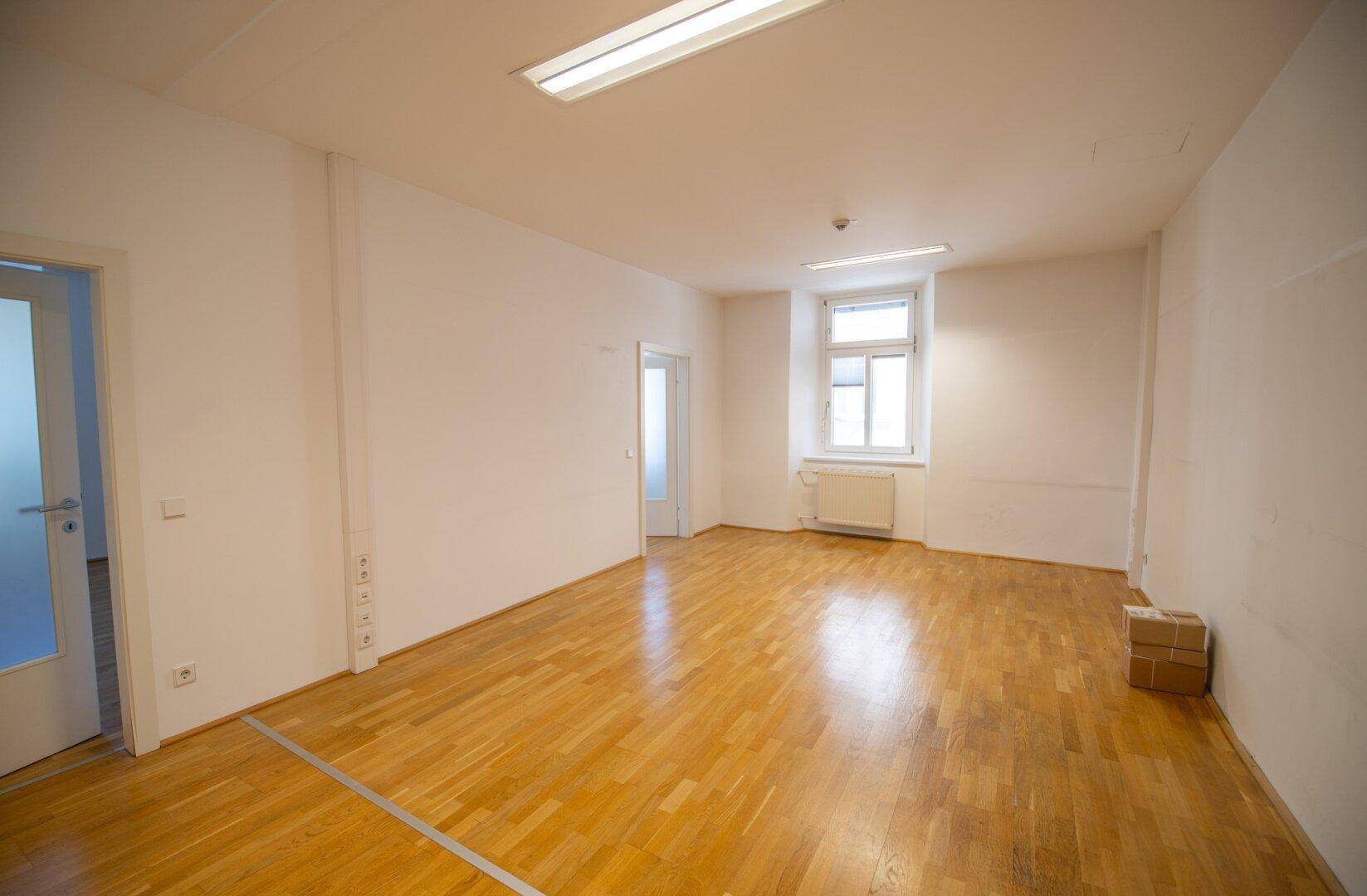 Geräumiger, heller Raum mit Parkettboden, weißen Wänden und einem Fenster.