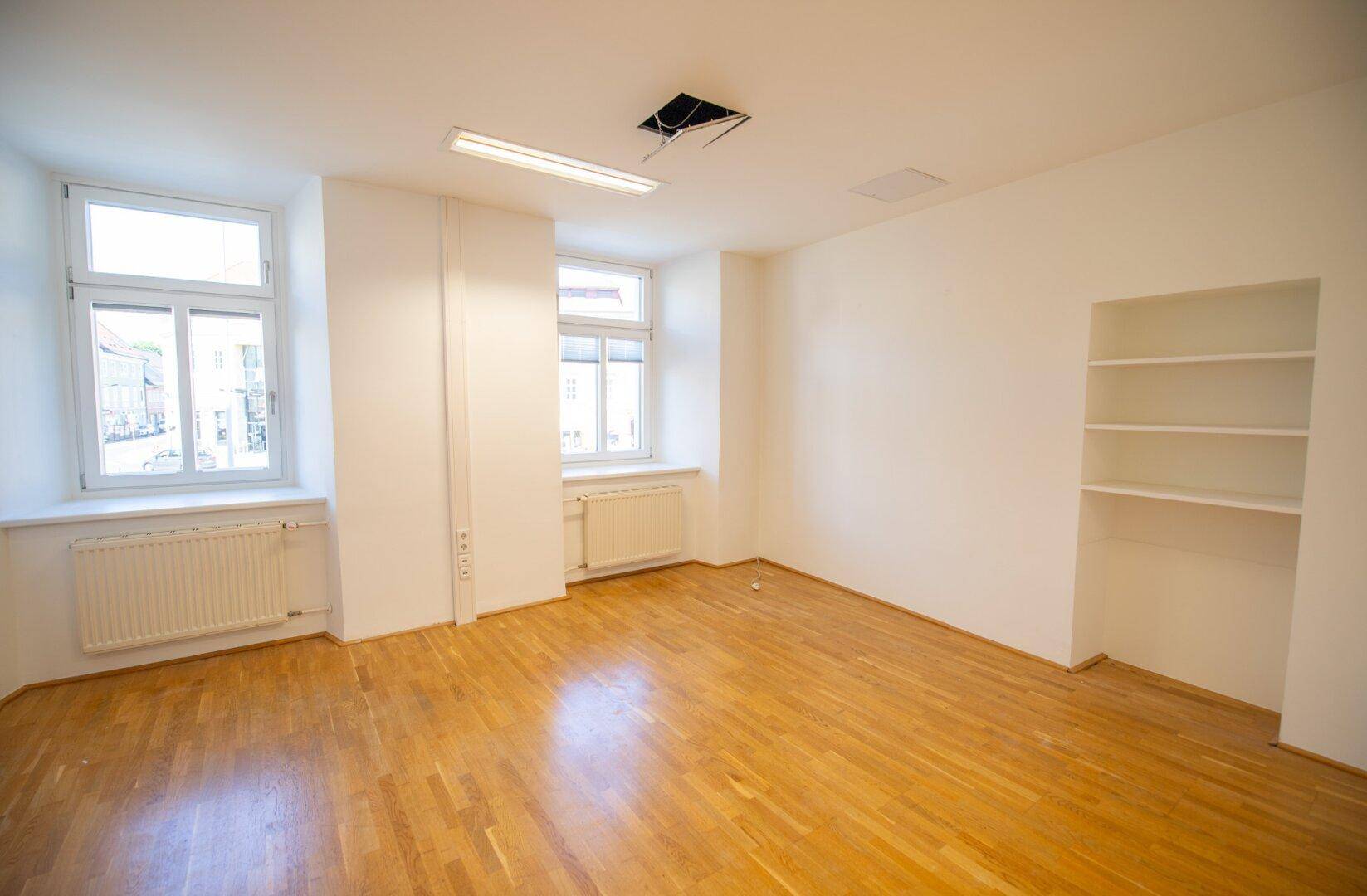 Heller Raum mit Parkettboden, weißen Wänden, zwei Fenstern und einer eingebauten Regalwand.