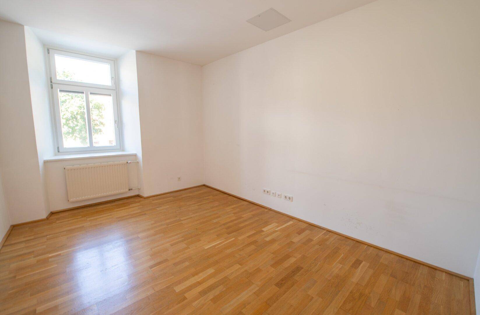 Einfacher, heller Raum mit Parkettboden, weißen Wänden und einem einzelnen Fenster.