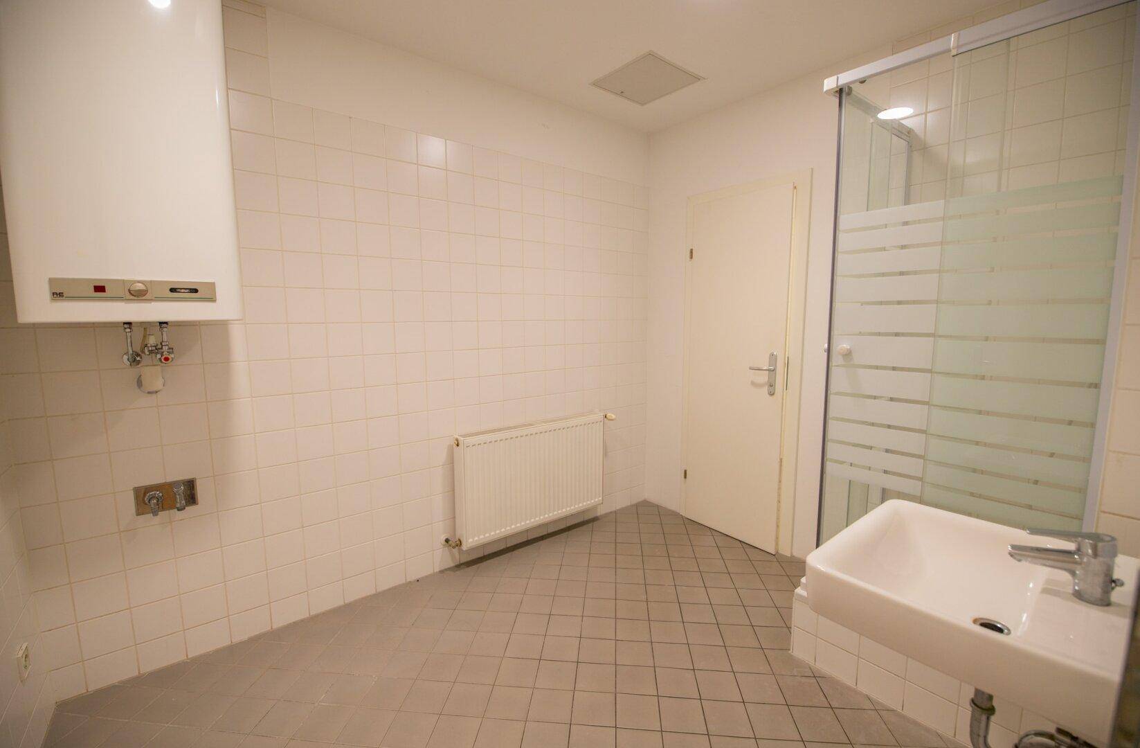 Badezimmer mit hellen Fliesen, Duschkabine, Waschbecken und Warmwasserbereiter.