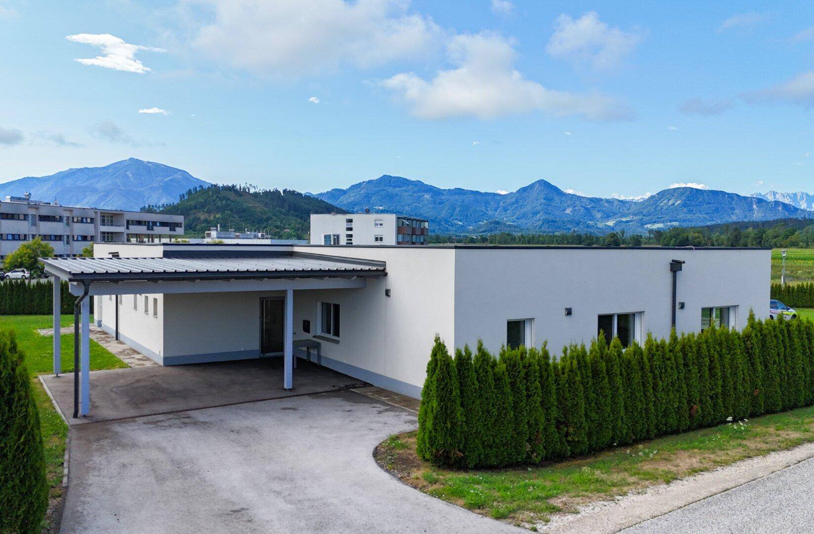 Modernes Gebäude mit Carport und gepflegter Hecke, im Hintergrund eine beeindruckende Berglandschaft.